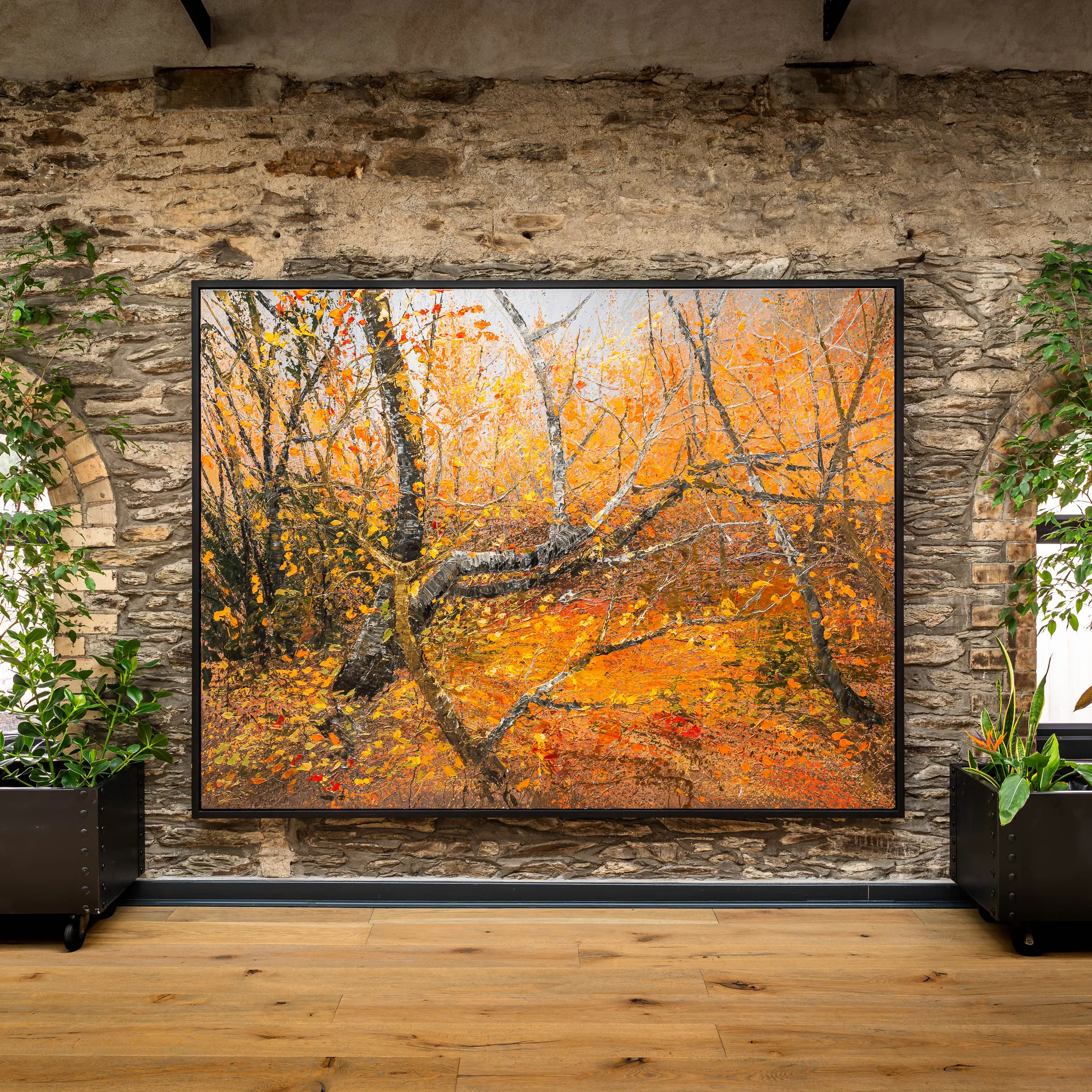 Autumn Leaves, an original painting by Paul Kenton, hangs in a black frame on a stone wall above a wooden floor. Its bold orange and gold trees and fallen leaves glow in a dense autumn wood.