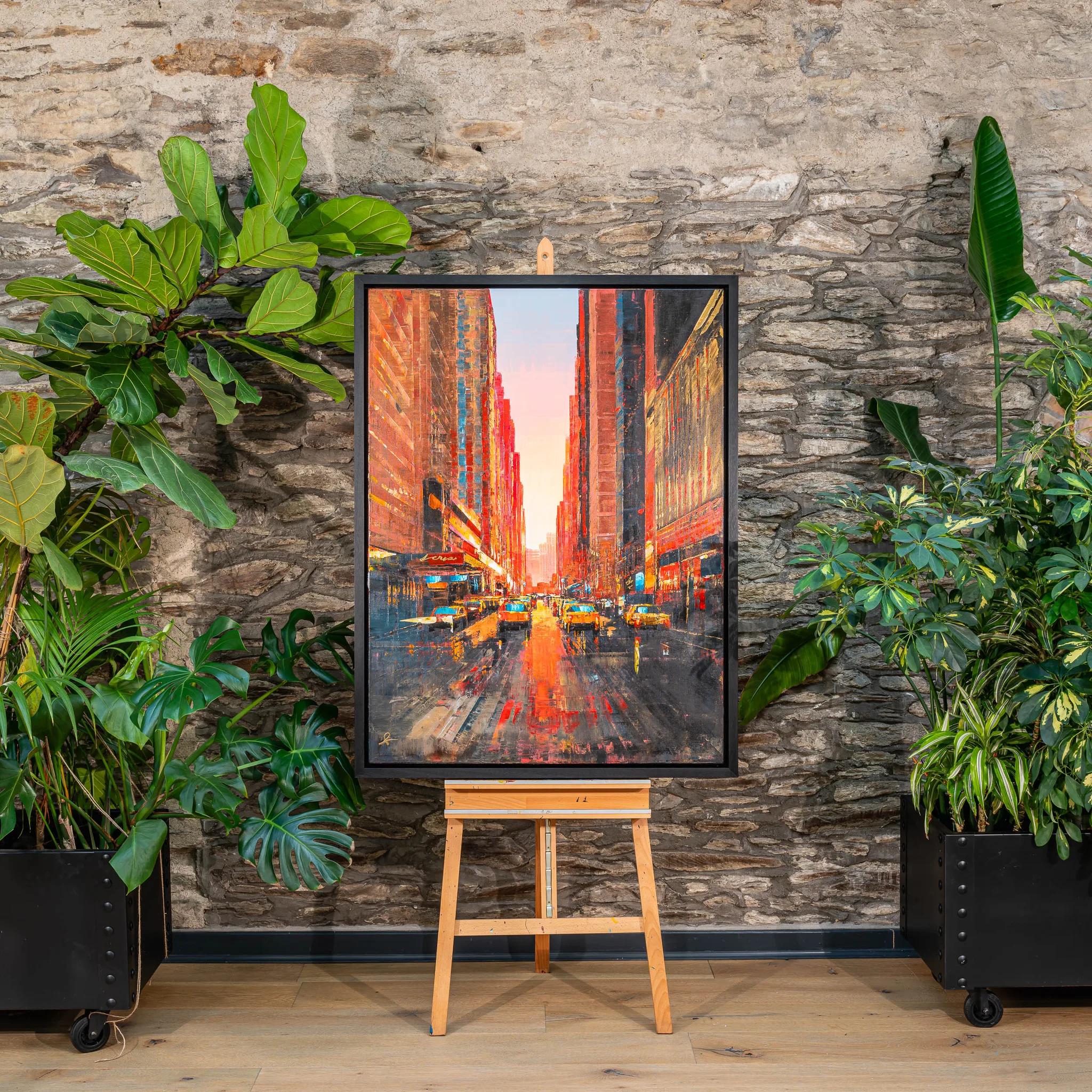 Big Apple Dreaming, an original painting by Paul Kenton, framed on an easel before a stone wall and tall plants. A sunset city street of yellow taxis and glowing towers recedes beneath a pale sky.