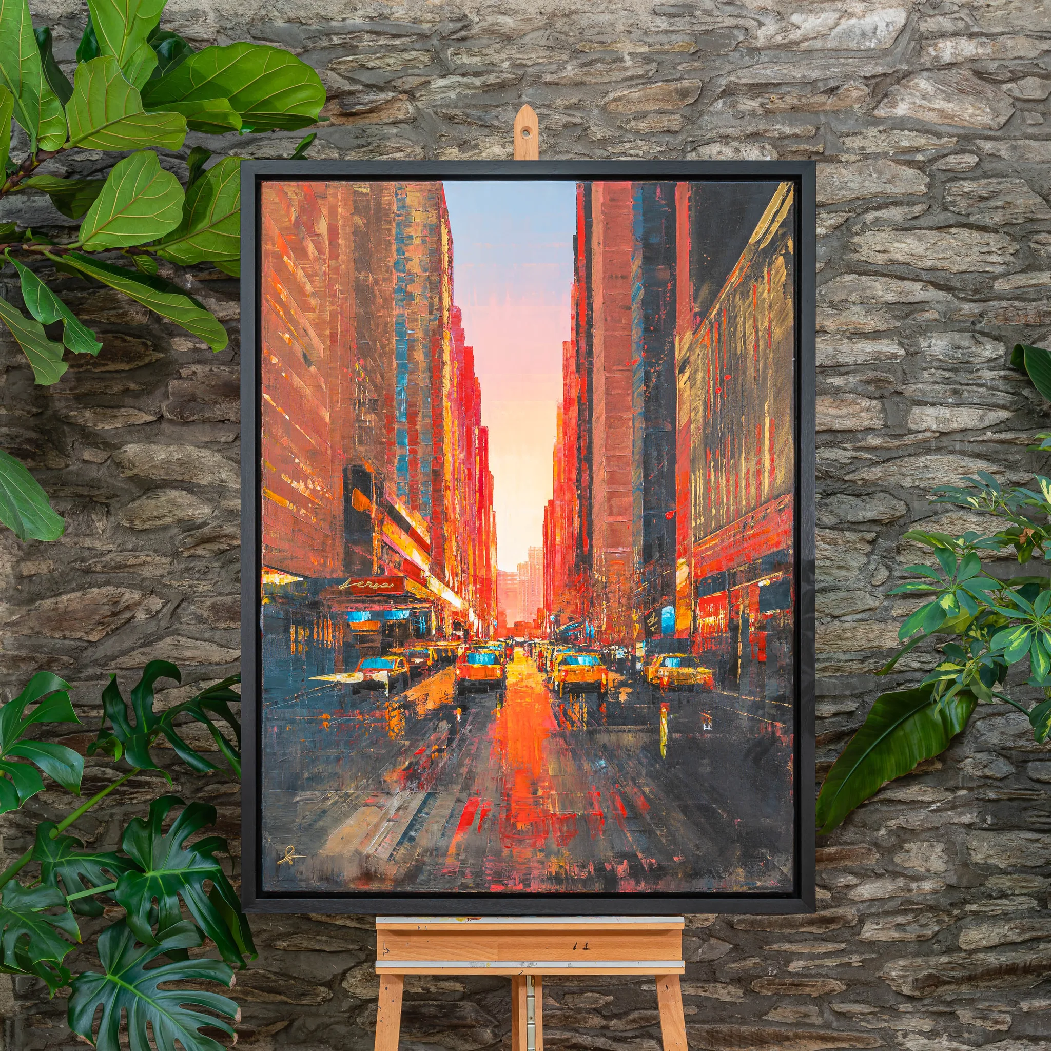 Big Apple Dreaming, an original painting by Paul Kenton in a black frame on an easel against a stone wall with leafy plants. A sunset street of New York, yellow taxis glowing amid orange and red towers.