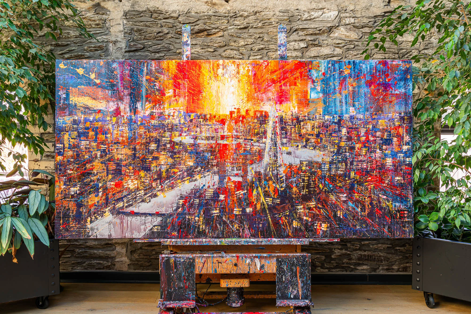 Blaze Across the Thames, an original mixed media painting by Paul Kenton, hangs on an easel before a stone wall, its bold fiery orange and red London skyline glowing above a dense mass of lit windows.