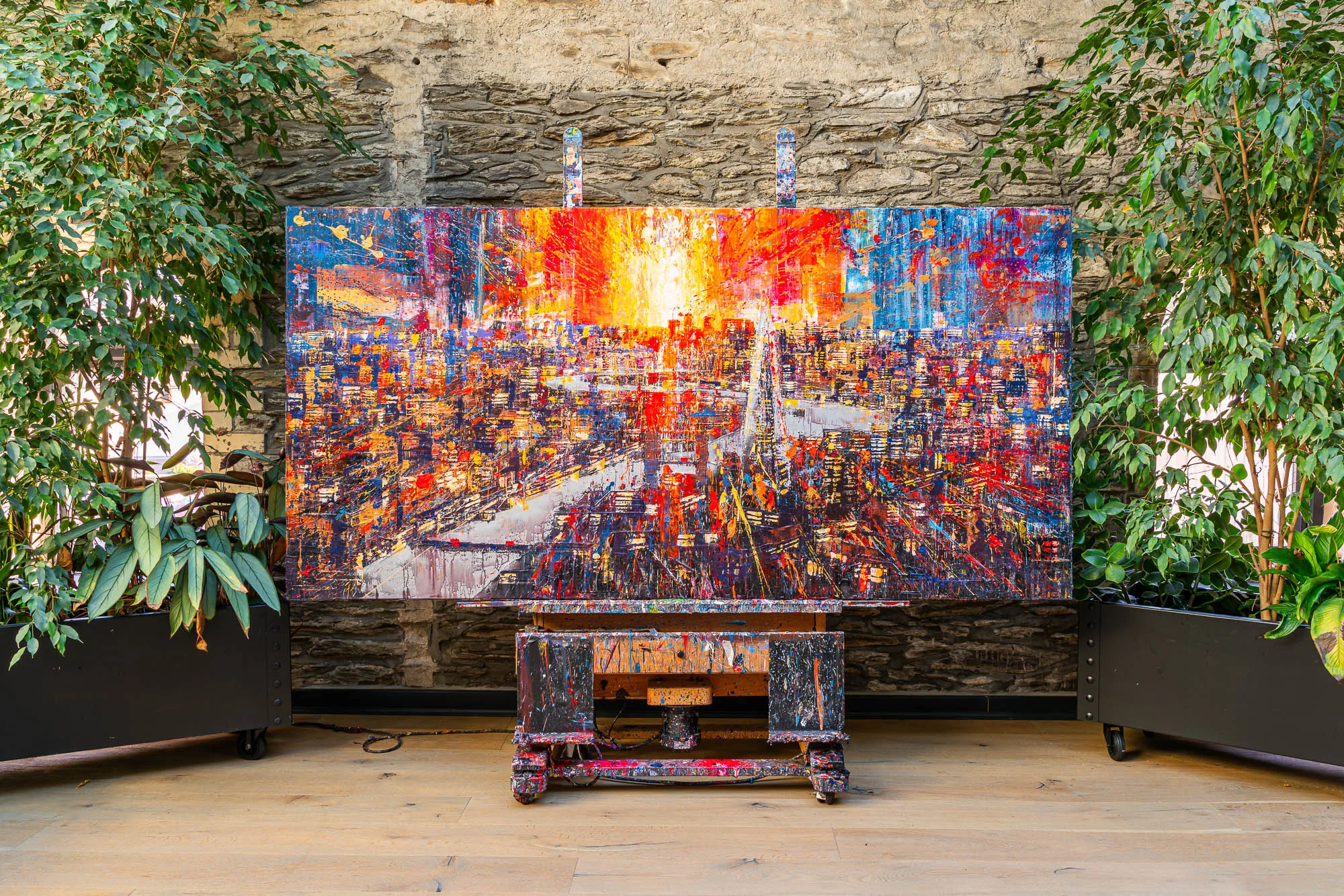 Blaze Across the Thames, an original painting by Paul Kenton, shown on an easel in a room with stone walls and plants, its textured London skyline glowing orange and red beneath a fiery sunset.