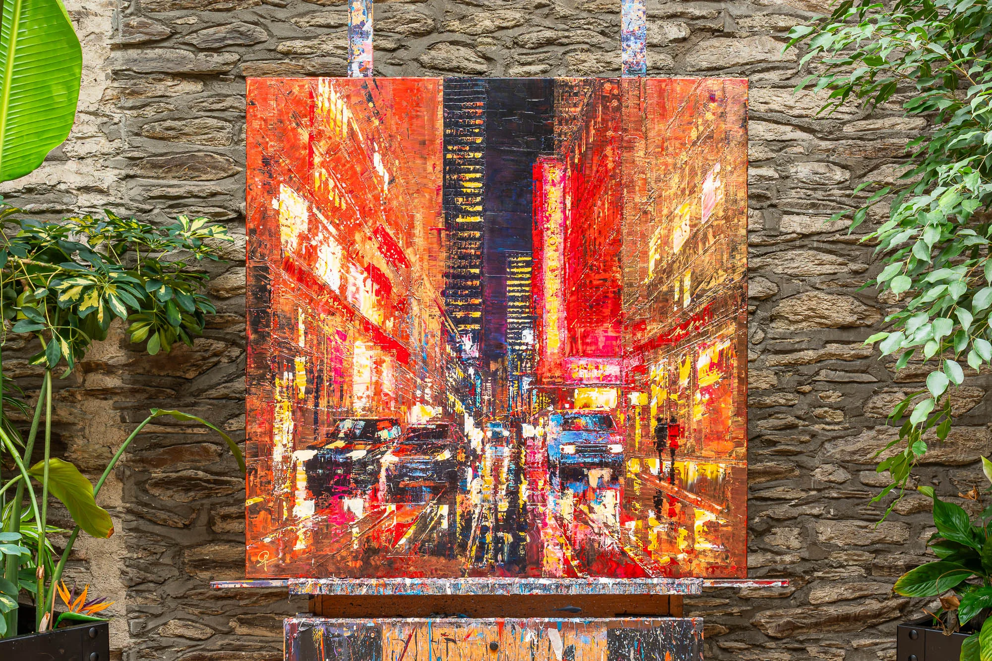 Blazing Broadway, an original mixed media painting by Paul Kenton, hangs on an easel against a stone wall between green plants. It shows a night street of glowing red and orange city lights, with cars and a figure in the glare.