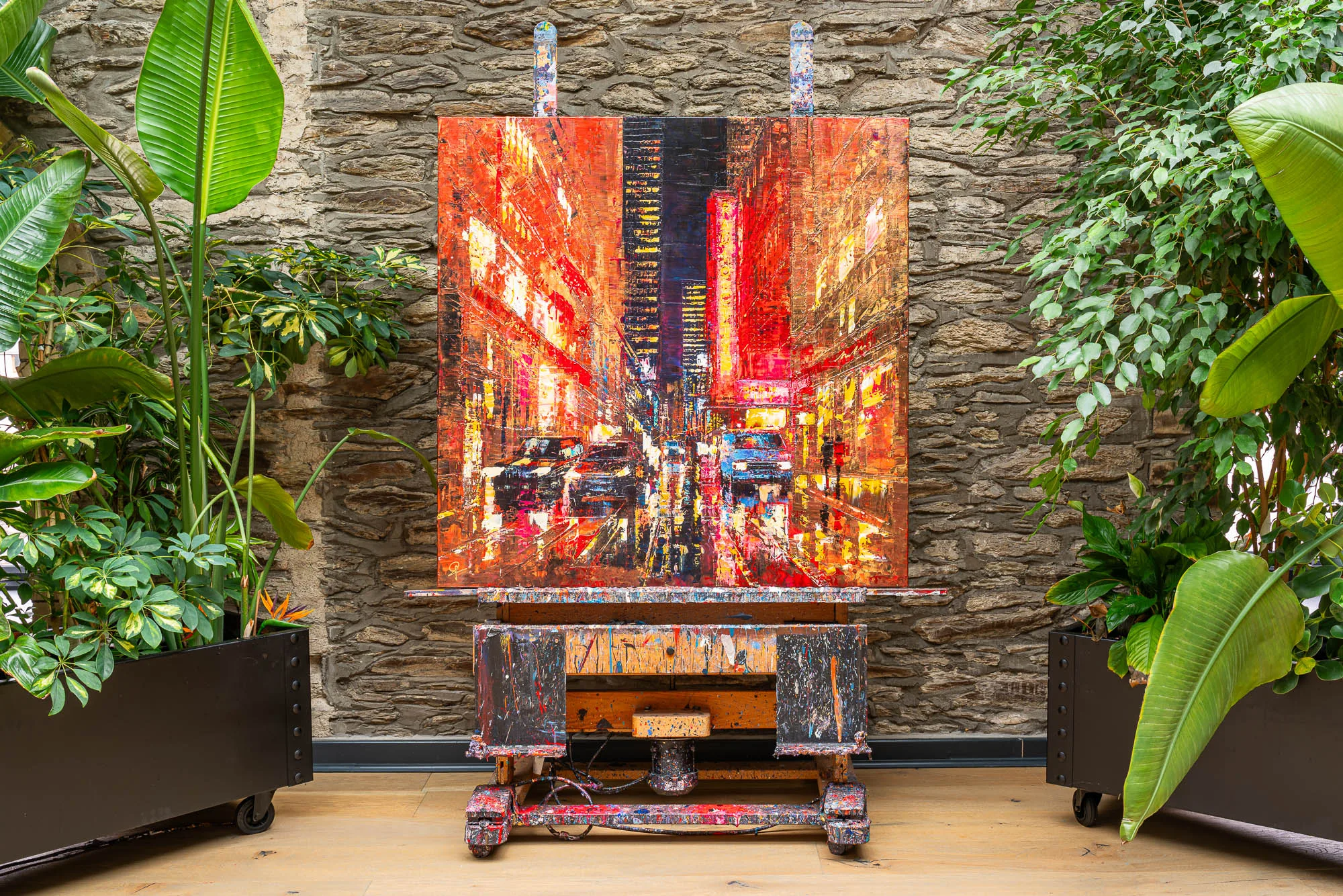 Blazing Broadway, an original painting by Paul Kenton, hangs on a stone wall between leafy plants, its glowing red and gold New York street scene framed by a paint-splattered wooden easel.
