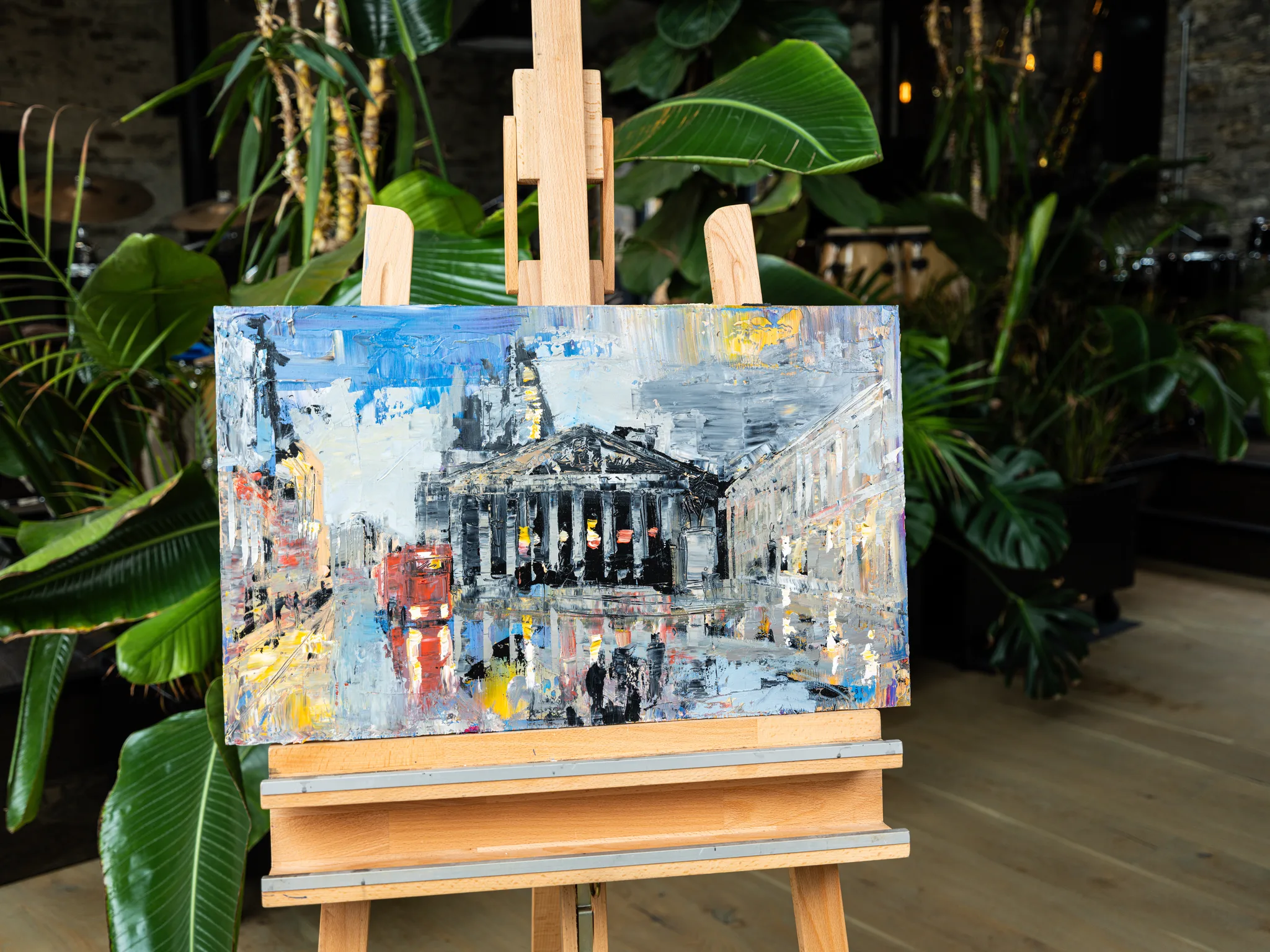 Bank Junction, an original painting by Paul Kenton on a wooden easel among large green plants: a textured London street, grey-blue with a red bus.