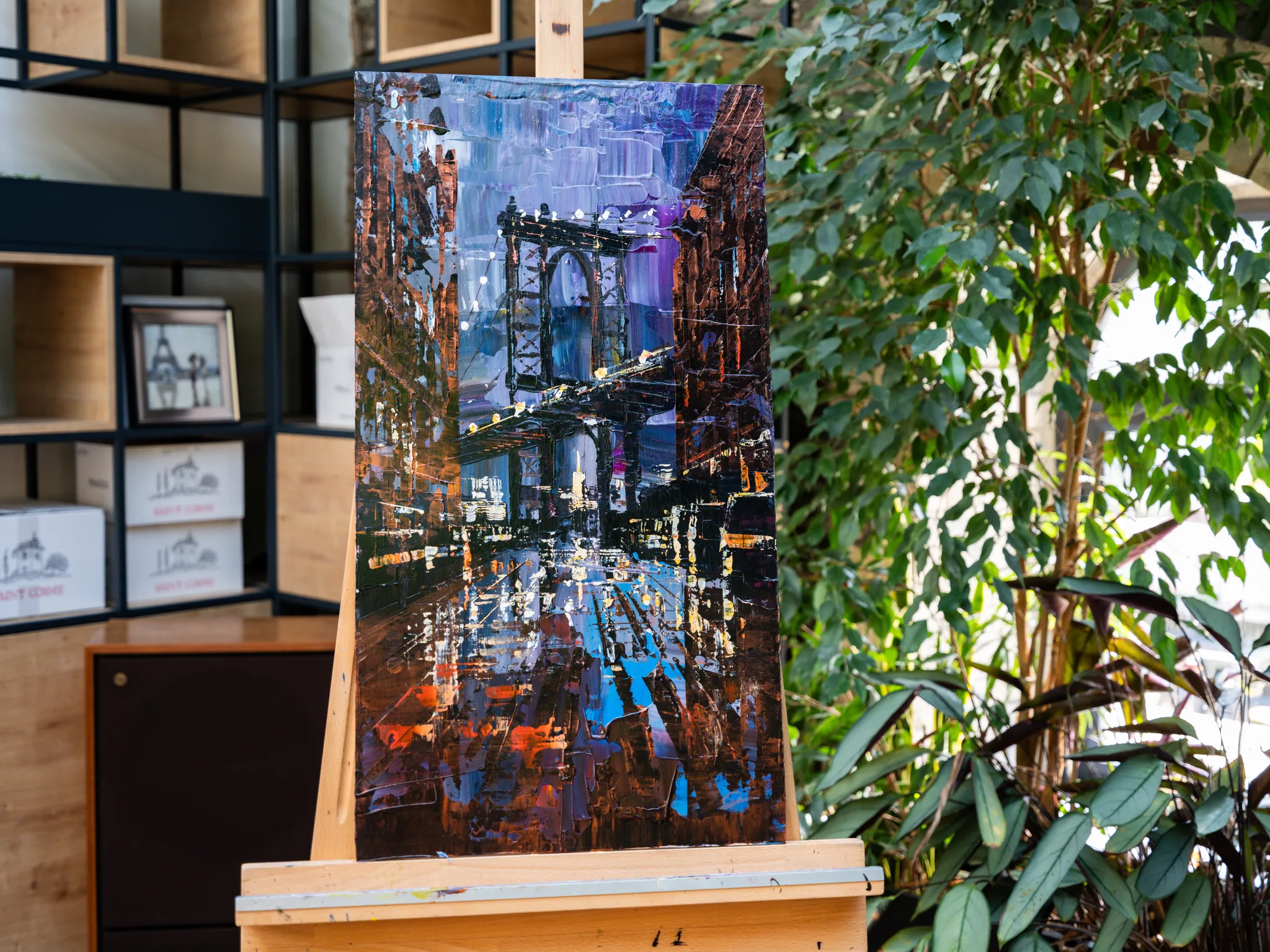 Bridge Nocturne, an original painting by Paul Kenton, on a wooden easel among green plants. A night view of a New York street framed by a bridge's arch, in blues, purples and warm orange lights.
