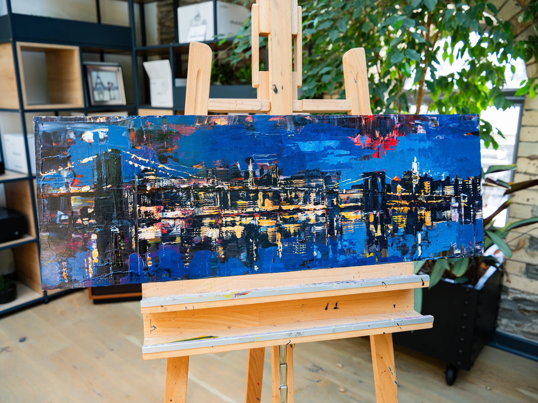 City After Dark, an original painting by Paul Kenton on an easel in a bright room: a long horizontal New York skyline at night, blue sky, glowing yellow and orange lights reflected in dark water.