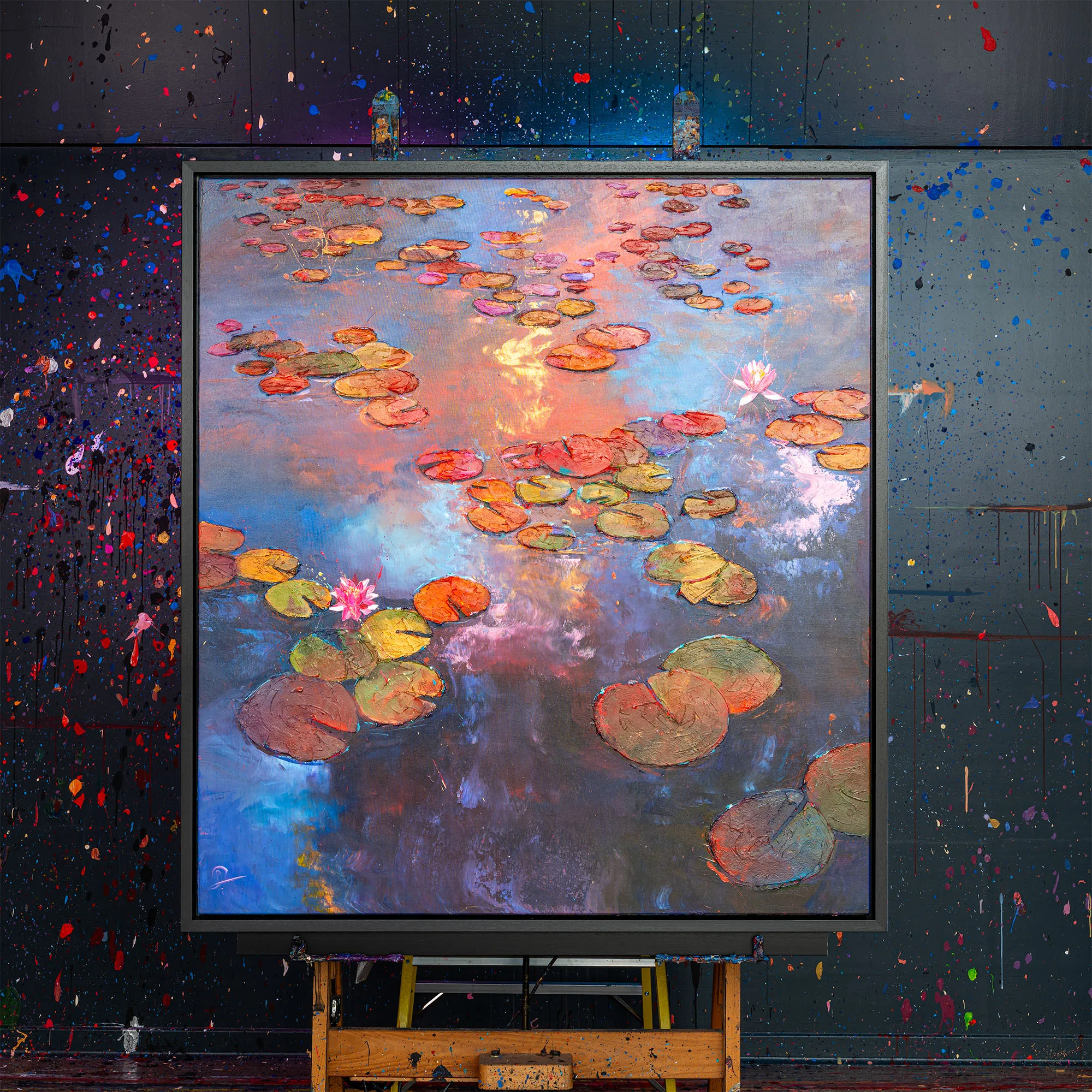 Celestial Blooms, an original painting by Paul Kenton, hangs in a dark studio splattered with paint. It shows lily pads and pink water lilies on a pond reflecting coral and blue sky.