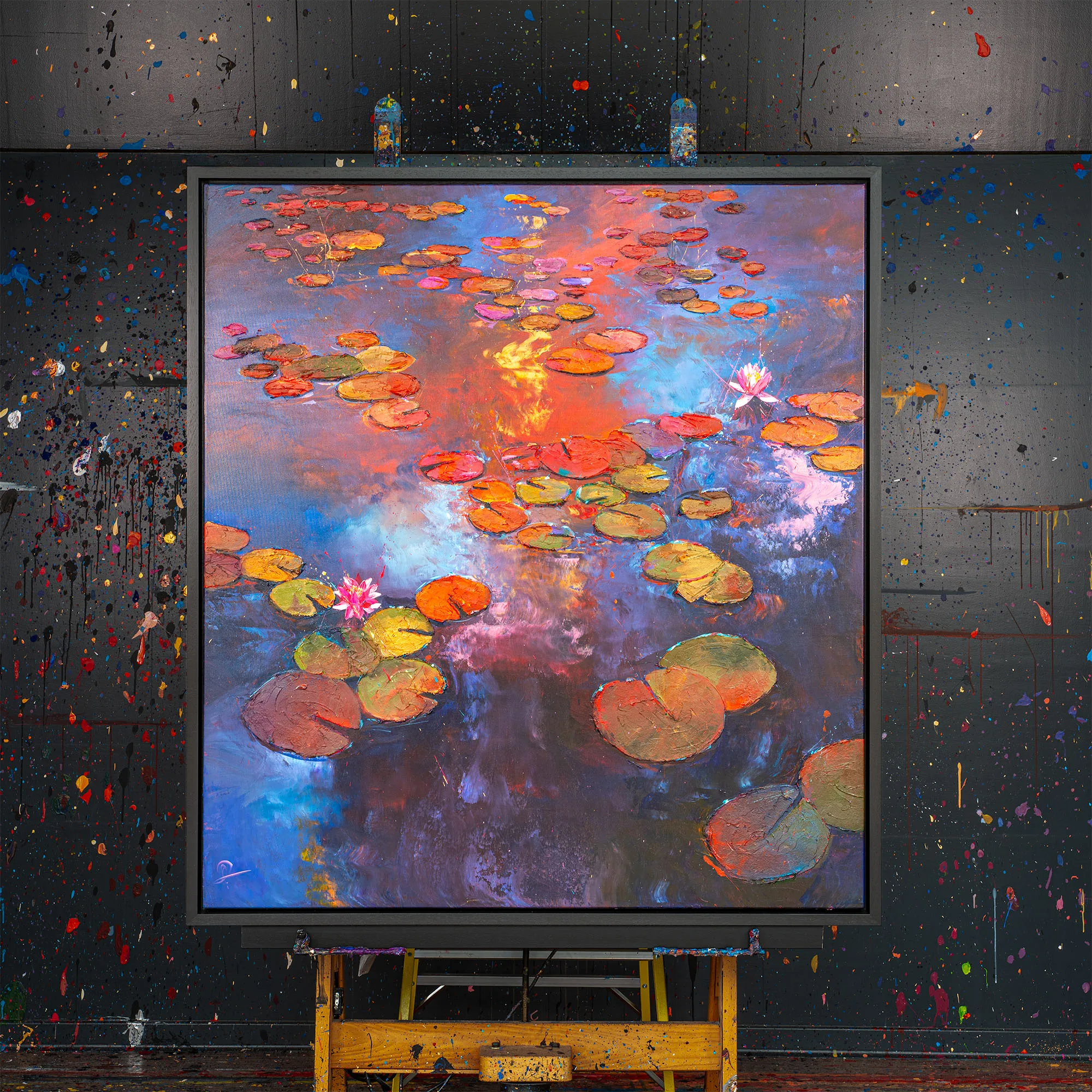 Celestial Blooms, an original painting by Paul Kenton, hangs in a dark studio splattered with paint, showing lily pads and pink blooms on a pond glowing orange and blue.