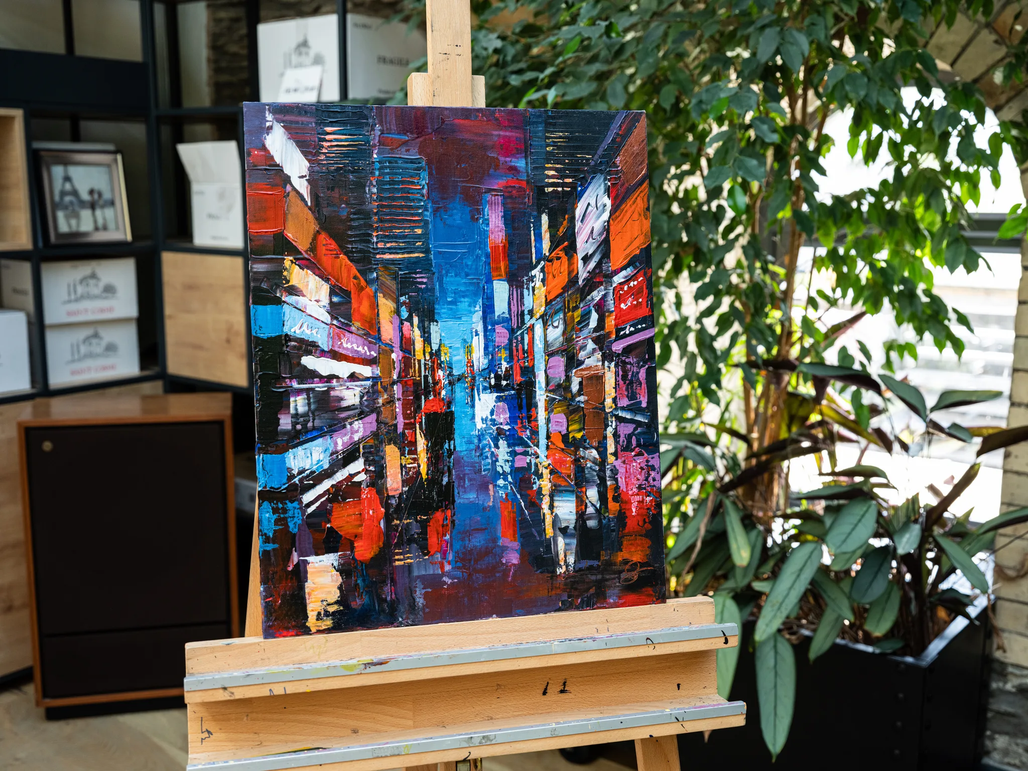 City Corridor, an original painting by Paul Kenton, displayed on a wooden easel in a shop. A narrow night street of glowing blue, red and orange shopfronts recedes towards a dark, reflective centre, in thick textured brushwork.
