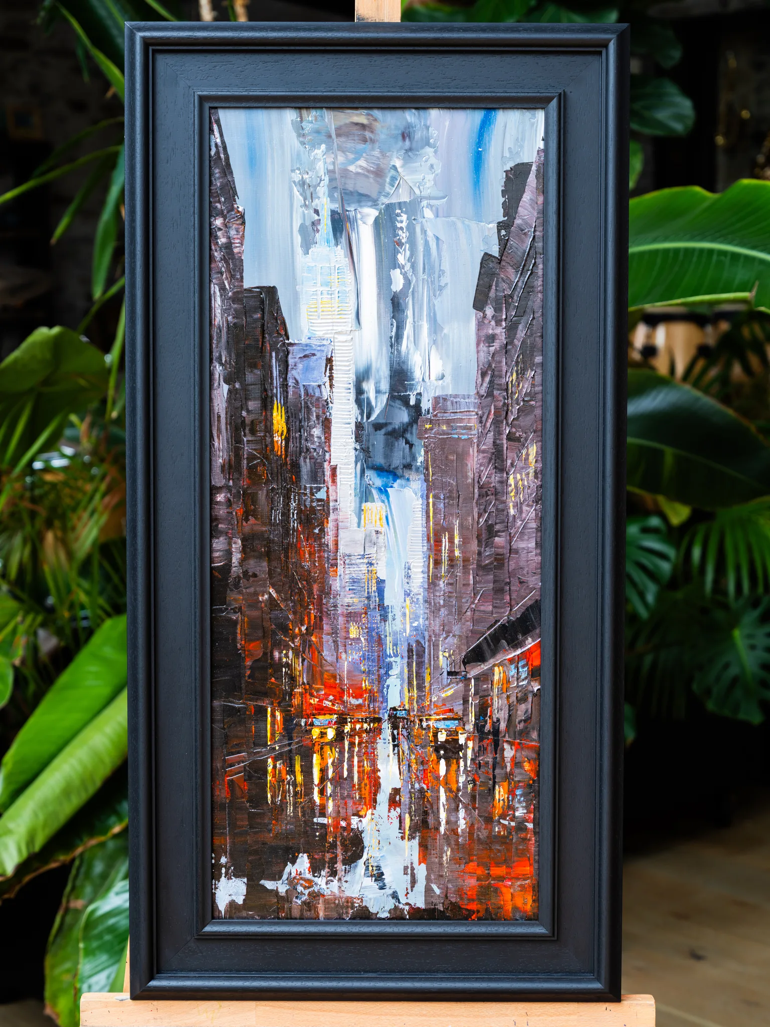 Dark Midtown, an original painting by Paul Kenton, shown in a black frame on an easel among green plants: a narrow New York street with towers under a pale sky, lit by warm orange reflections.