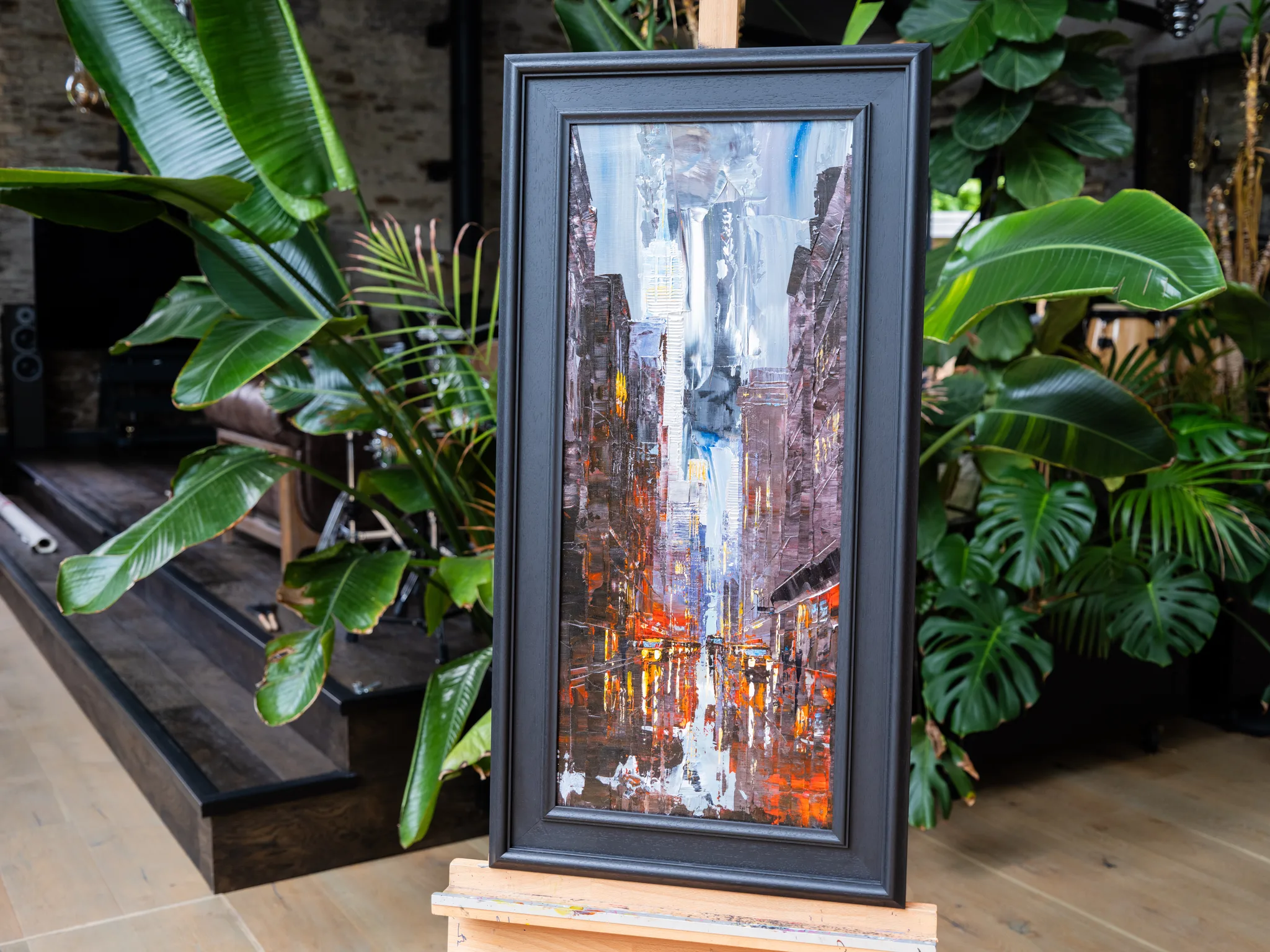 Dark Midtown, an original painting in a black frame on an easel among green plants, showing a dim New York street lit orange.