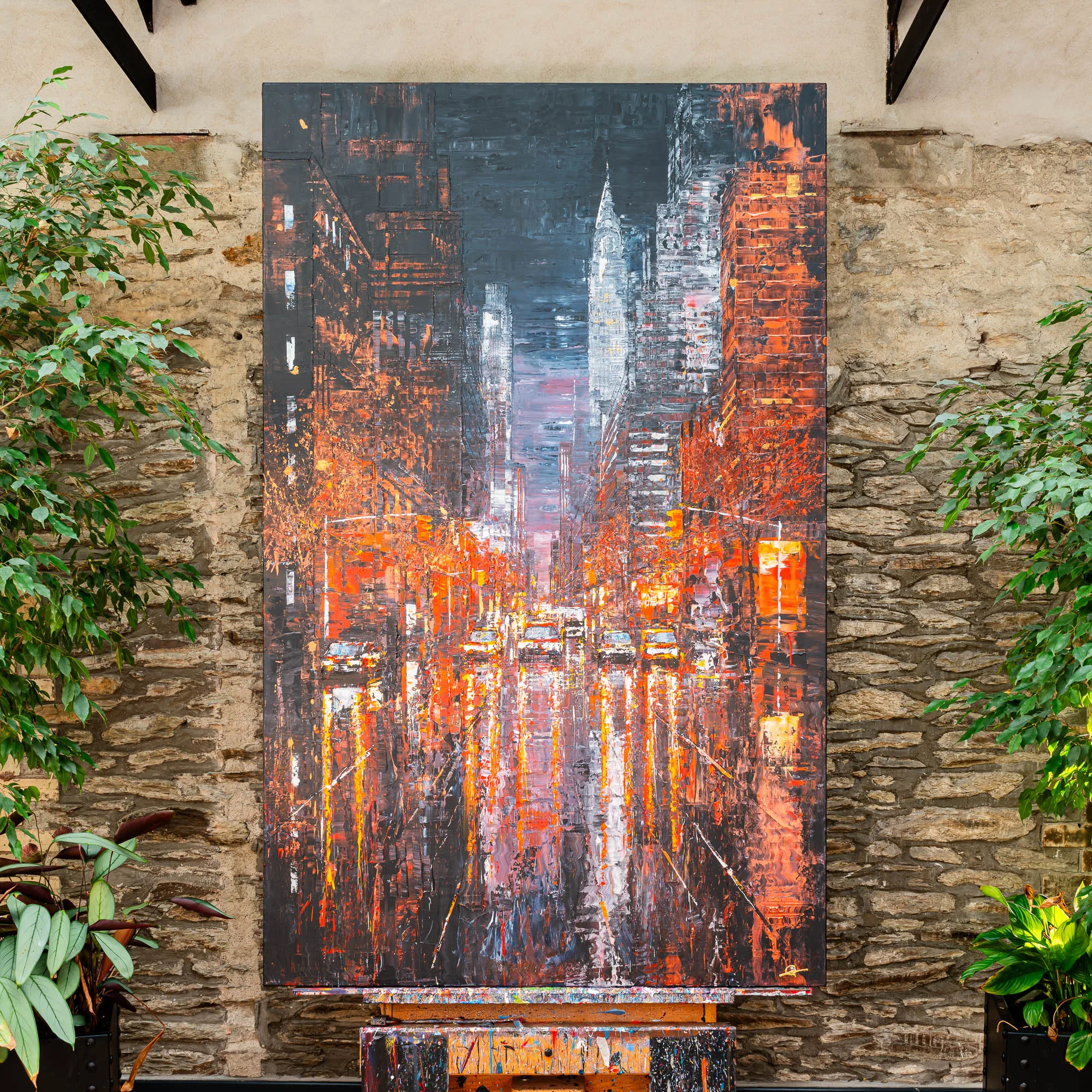Embers of the City, an original painting by Paul Kenton, hangs on a stone wall between green plants. It shows a night street of New York in fiery oranges and charcoal greys, with glowing reflections on wet road and thick textured brushwork.