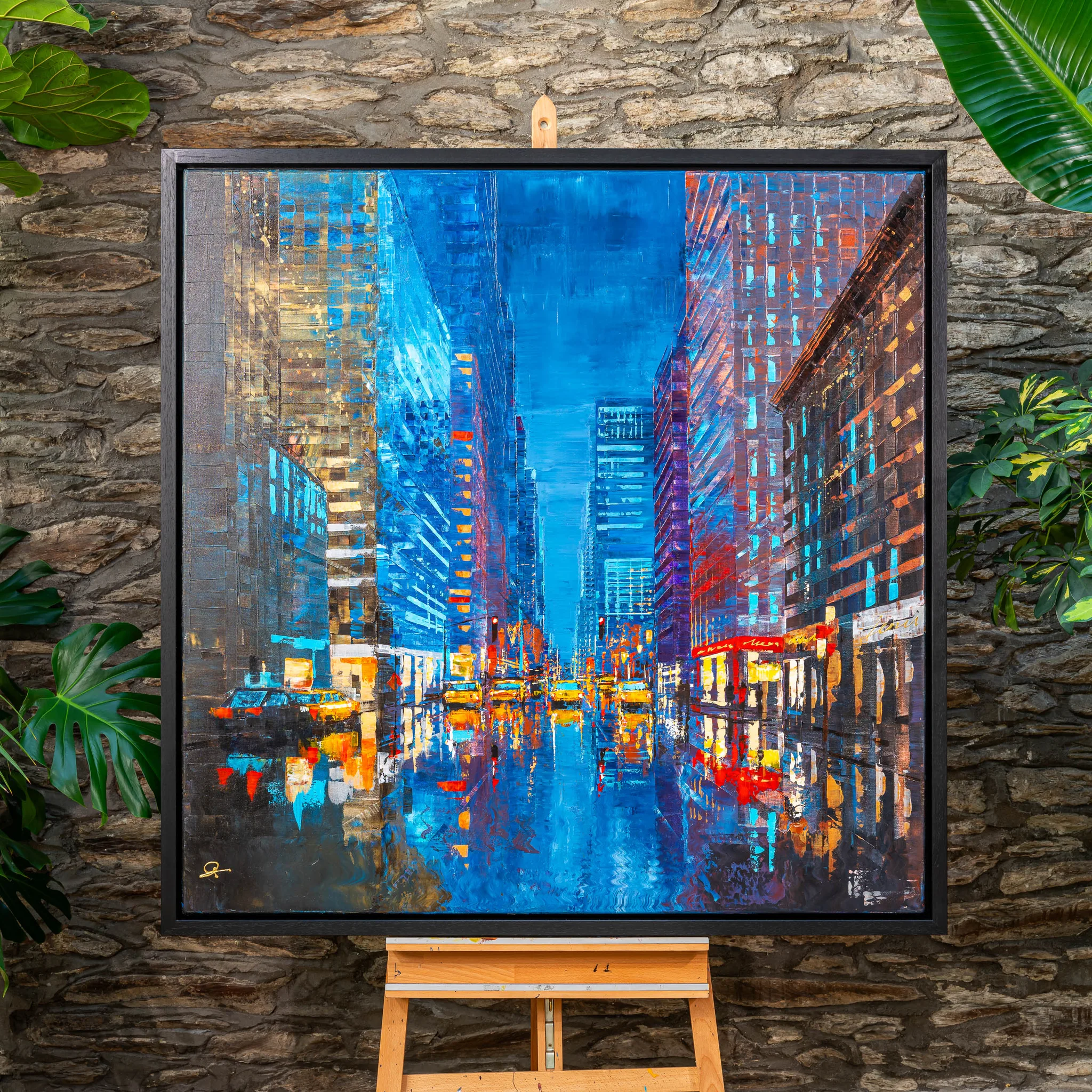 Feeling Groovy, an original painting by Paul Kenton in a black floating frame, hung on a stone wall beside green foliage and set on a wooden easel. It shows a bright blue city street at dusk, with yellow taxis and lit windows reflected in wet tarmac.