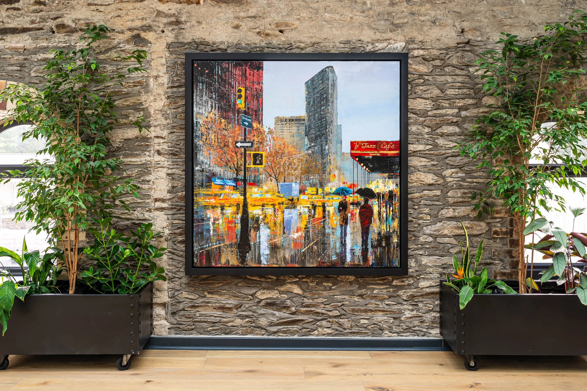 Flat Iron Hustle, an original painting by Paul Kenton, hangs in a black frame on a stone wall between two potted plants. It shows a rainy New York street of yellow taxis, umbrellas and reflections in red, orange and gold.