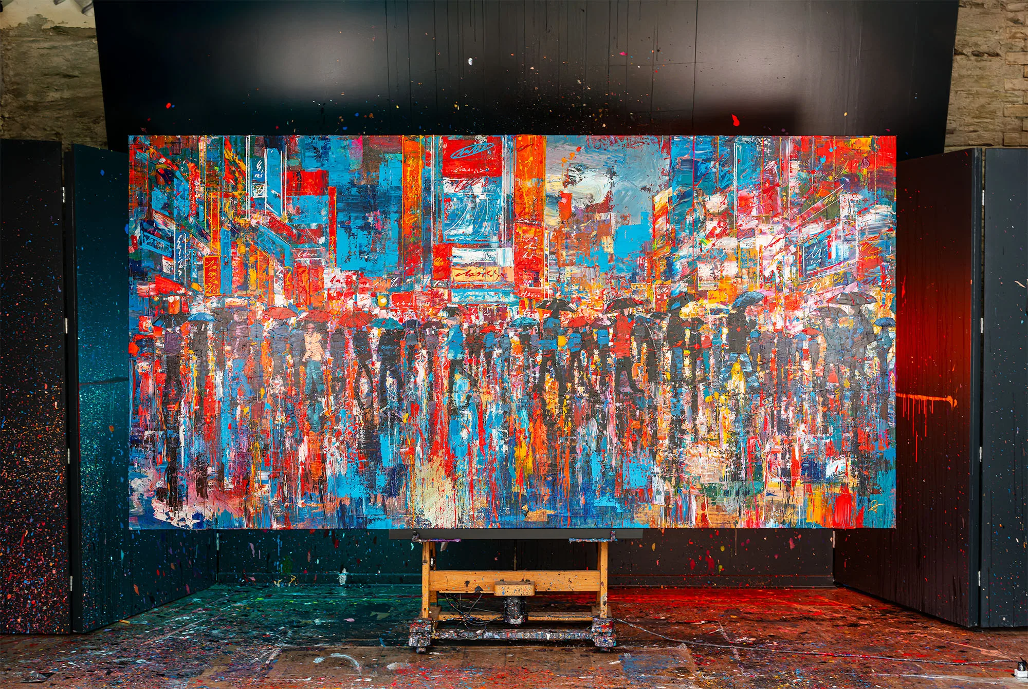 Fairytale of New York, an original mixed media painting by Paul Kenton, hangs on an easel in a studio splattered with paint. Its bold red, orange and blue city street, crowded with glowing signs and blurred figures, dominates the room.