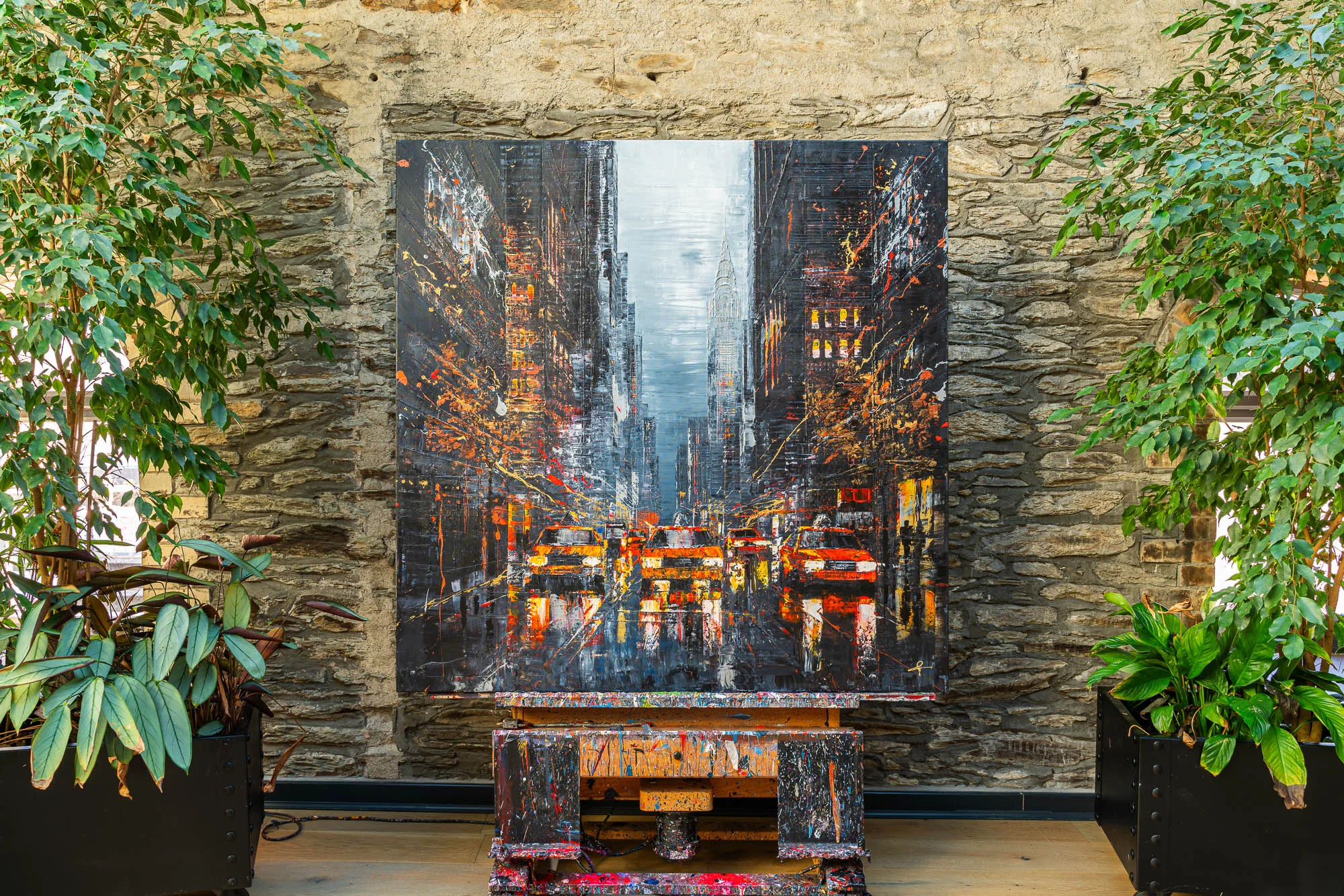 Flight of the Taxis, an original painting by Paul Kenton, hangs on a stone wall between leafy plants, showing a dark, splattered city street of glowing yellow and orange taxis under a pale sky.