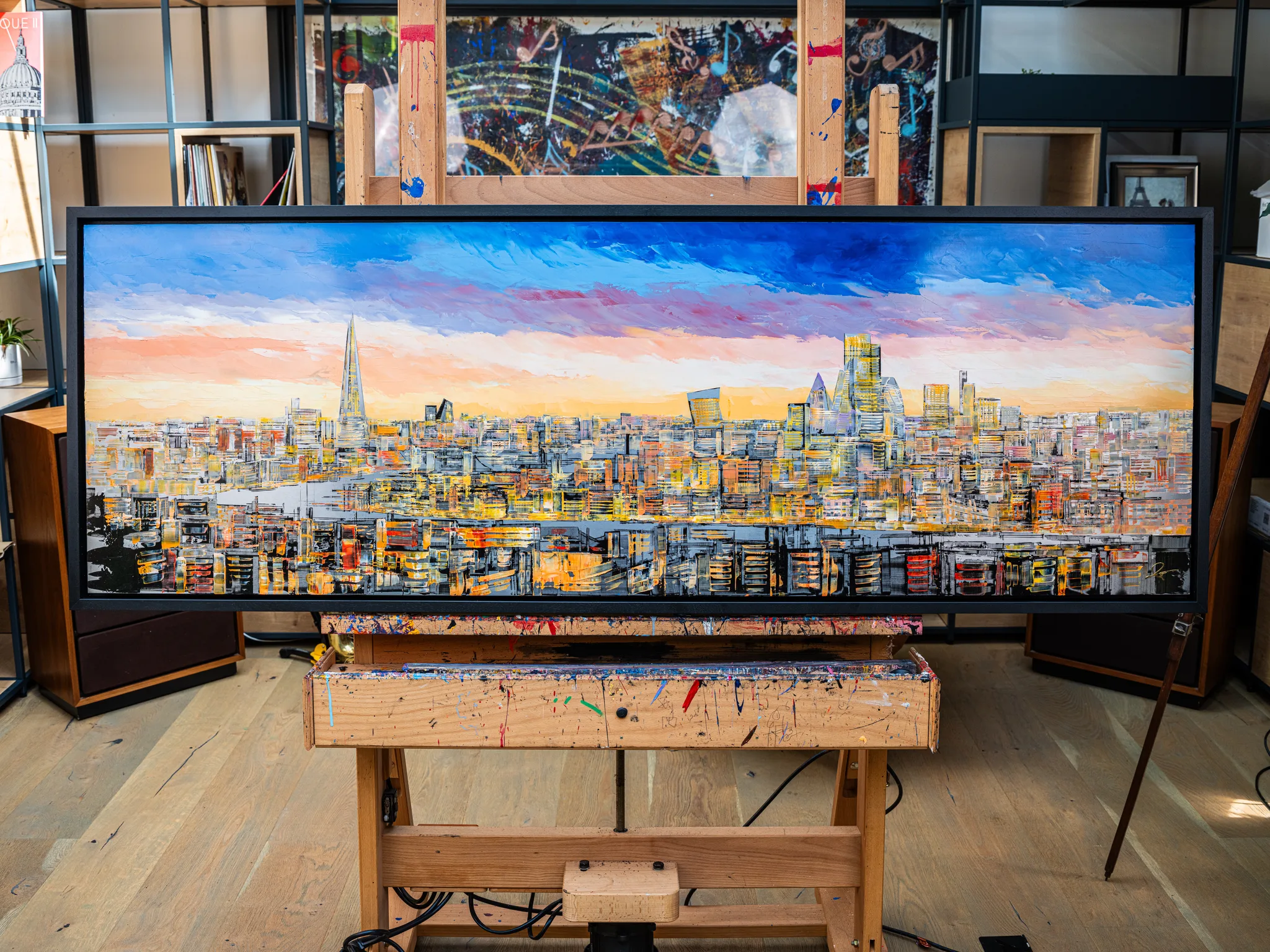 Golden Capital, an original painting by Paul Kenton, hangs in a dark frame in a studio. It shows a textured London skyline at dusk, with the Shard rising on the left under a blue and peach sky.