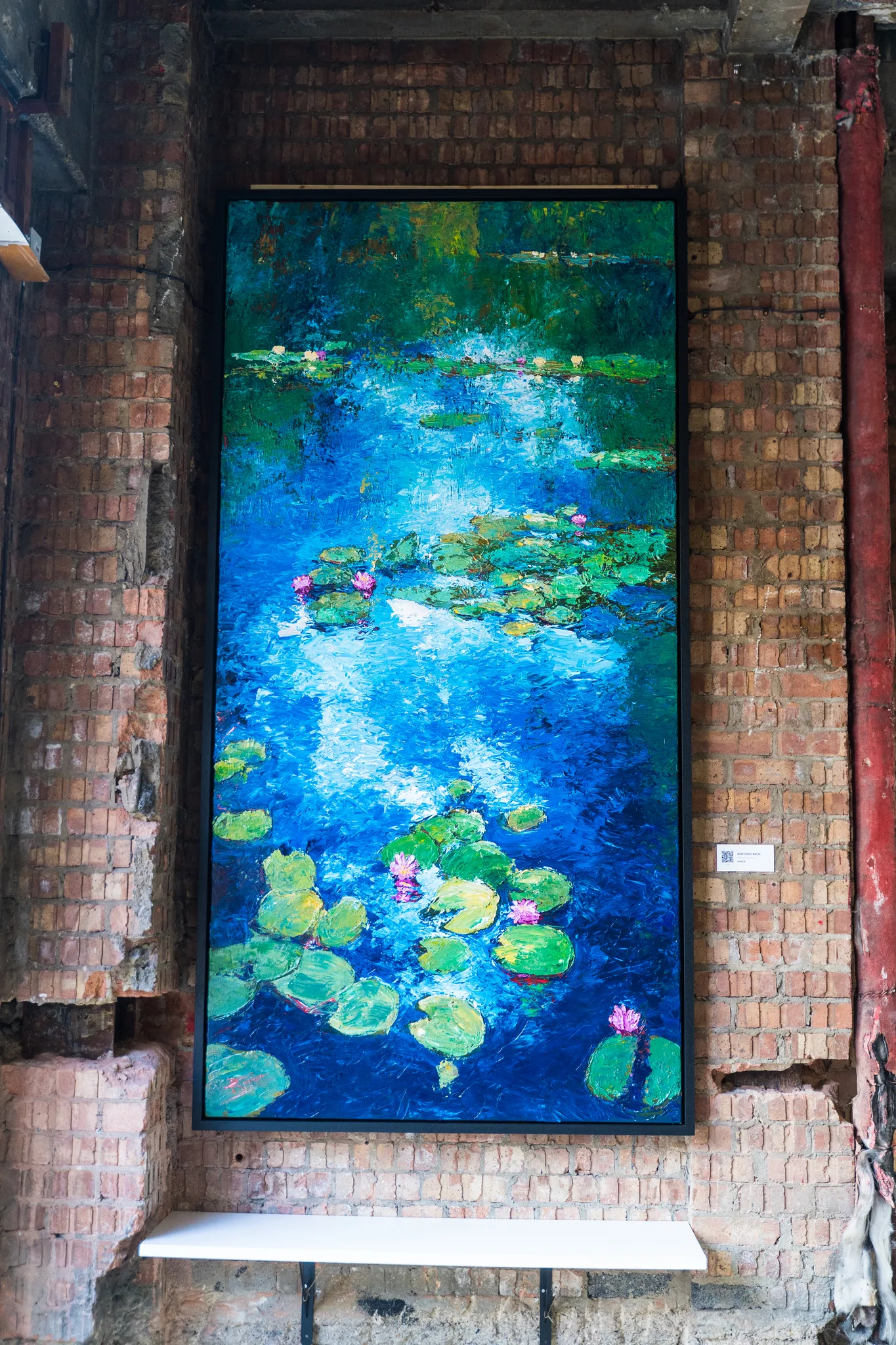 Impressionist Waters, an original painting by Paul Kenton hung on a brick wall: a tall canvas of a pond with pink water lilies and green lily pads over bold blue, textured water.