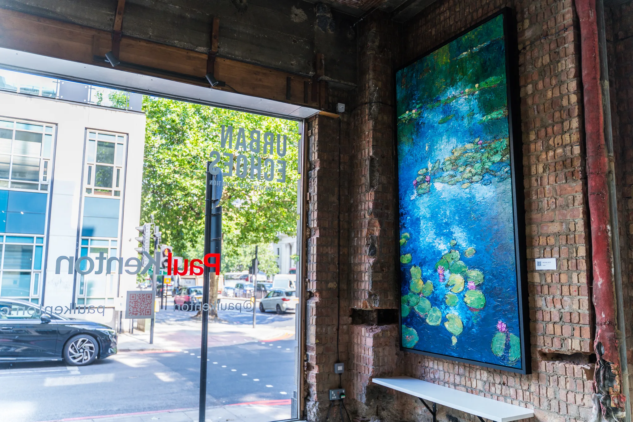 Impressionist Waters, an original mixed media painting by Paul Kenton, hangs on a brick wall in a shopfront window. It shows blue water with green lily pads and pink flowers, set beside a street view.