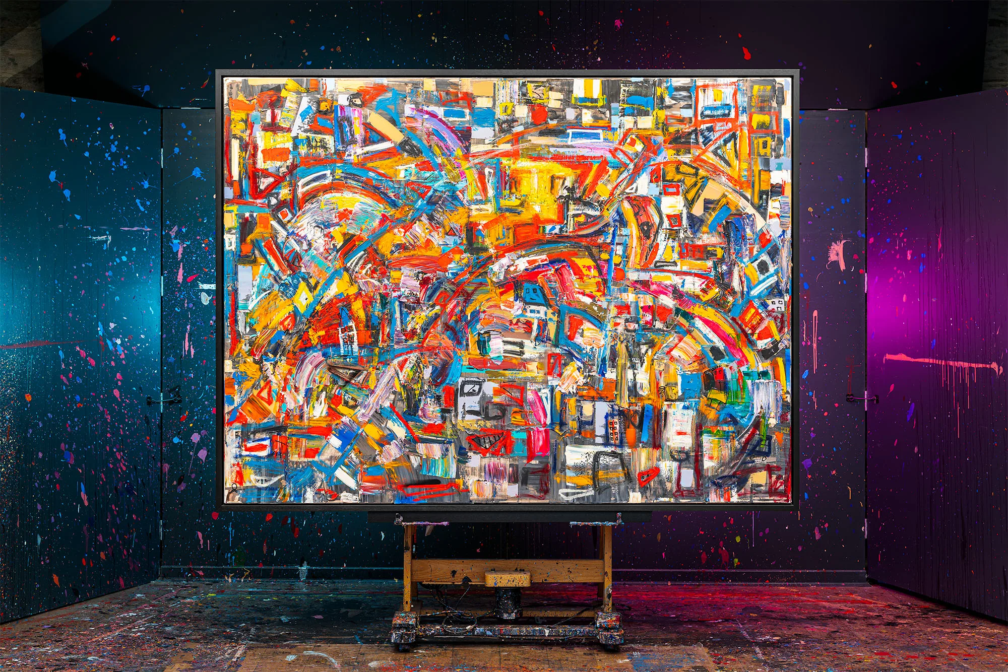 Laid Back, an original mixed media painting by Paul Kenton, hangs on an easel in a studio splashed with paint. Its busy, bold swirls of orange, red, yellow and blue read like a jostling city.
