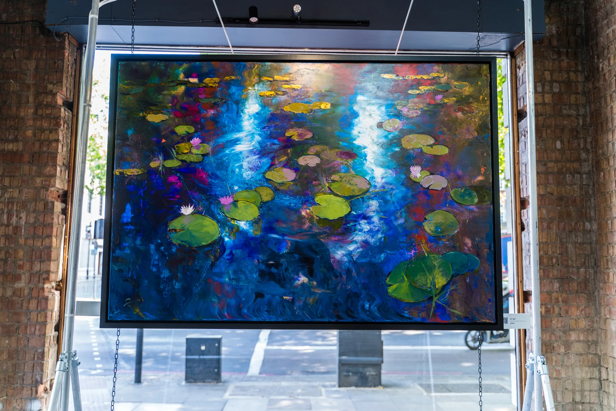 Lily Light, an original painting by Paul Kenton, hangs in a brick-walled gallery window above a street. Its deep blue water is flecked with pink, gold and green lily pads and blooms.