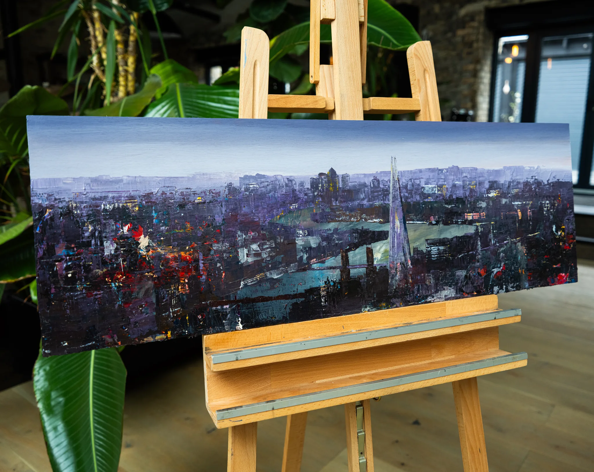 London Nocturne, an original mixed media painting by Paul Kenton, shown on a wooden easel in a room with plants. A wide panoramic London skyline at dusk in purples, greys and flecks of red and gold, with the Shard rising on the right over the river.