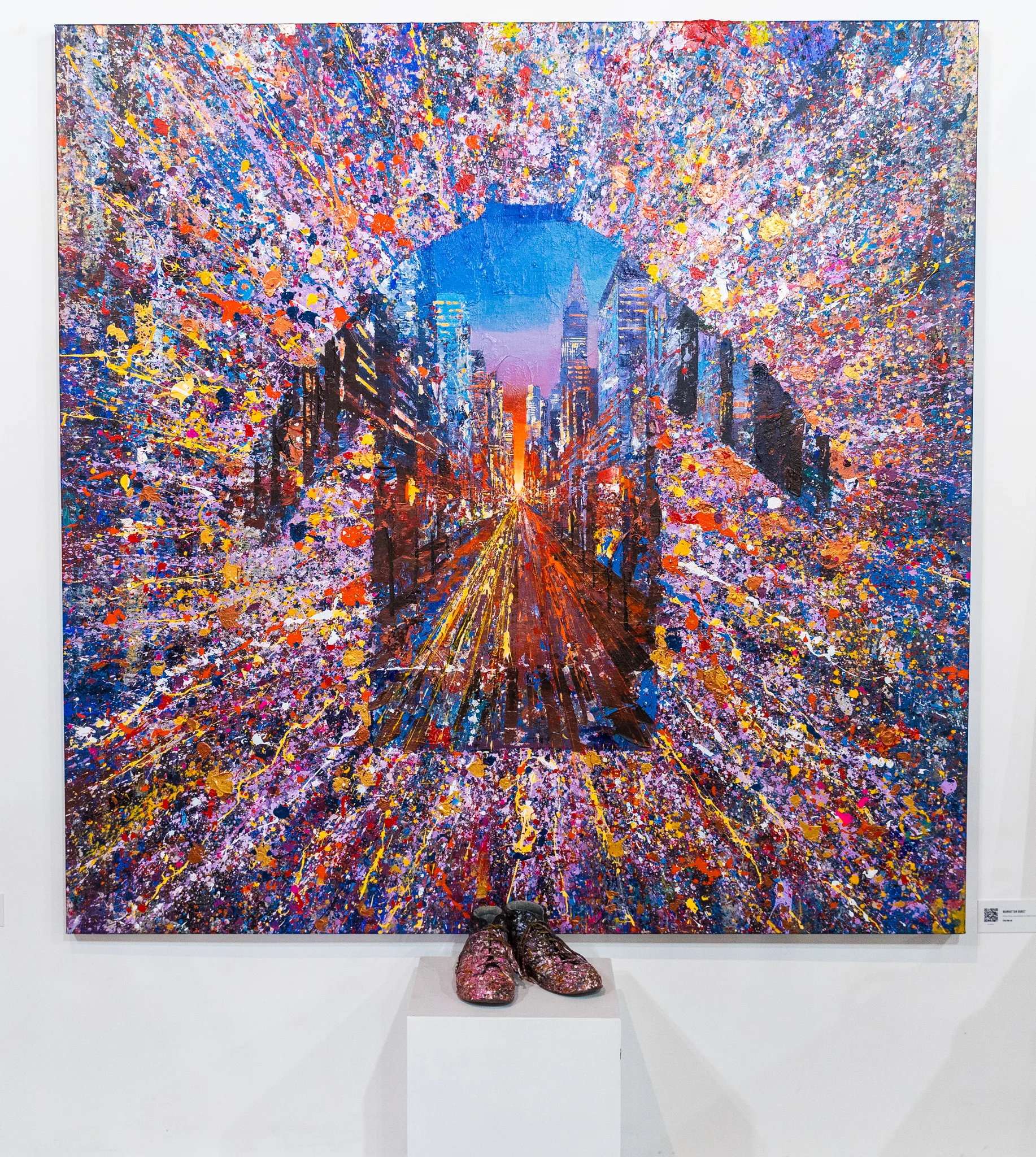 Manhattan Burst, an original painting by Paul Kenton, hangs on a white gallery wall. A central view of a city avenue at sunset is framed by bursts of multicoloured splatter, with a pair of glittering pink shoes on a plinth below.
