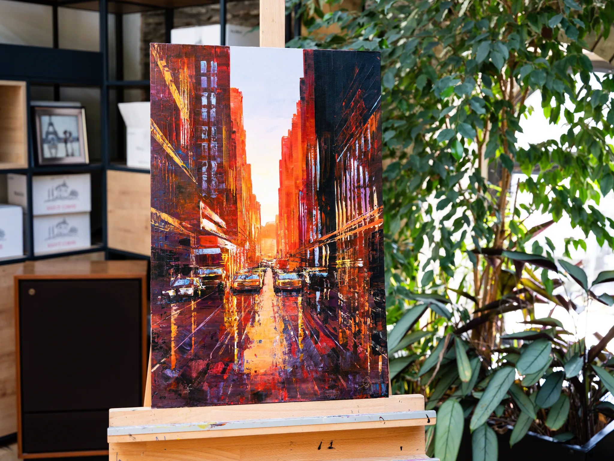 Manhattan Heat, an original painting by Paul Kenton, on a wooden easel in a bright room with plants. A sunset street of tall buildings glows orange and red, with yellow taxis and reflections on the wet road.