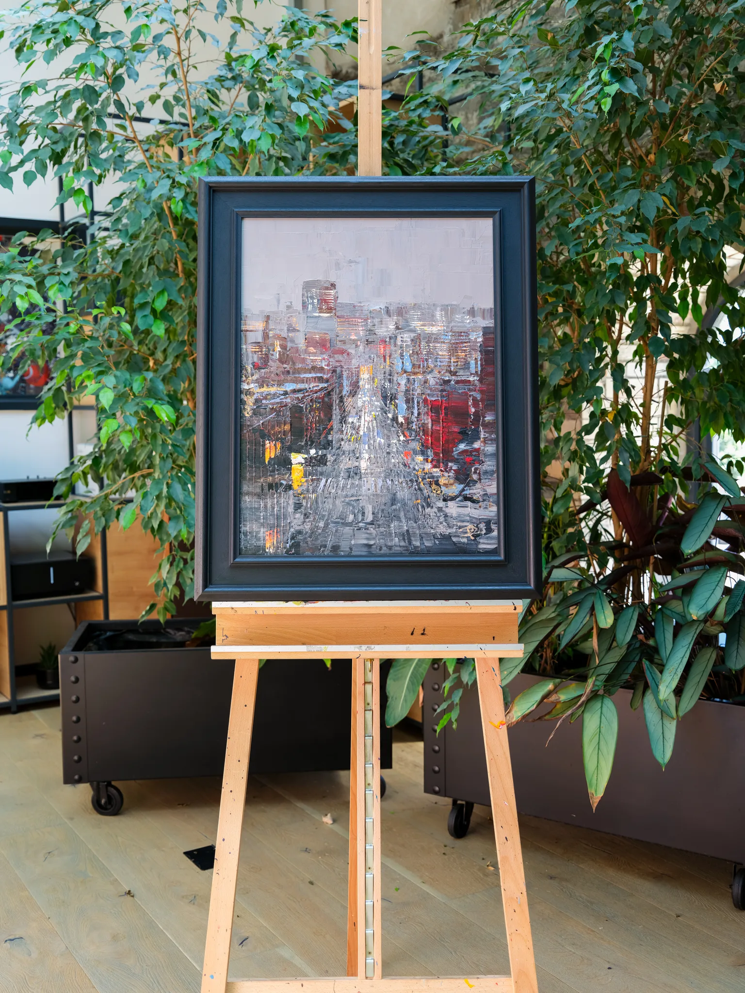 Midtown Silver, an original mixed media painting by Paul Kenton, framed in black on a wooden easel among indoor plants. A textured street view of New York in silver, grey and red, with tall buildings receding to a pale sky.