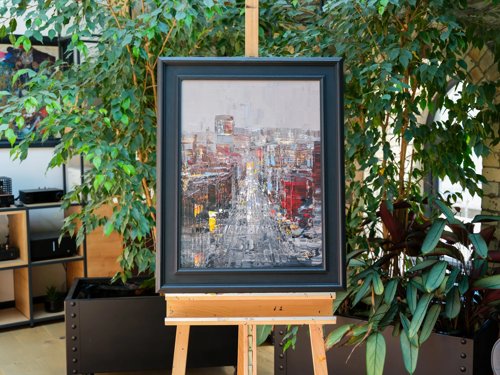 Midtown Silver, an original painting by Paul Kenton, shown in a dark frame on a wooden easel among leafy plants. A textured street view of New York in silver, grey and red, with tall buildings receding toward a pale sky.