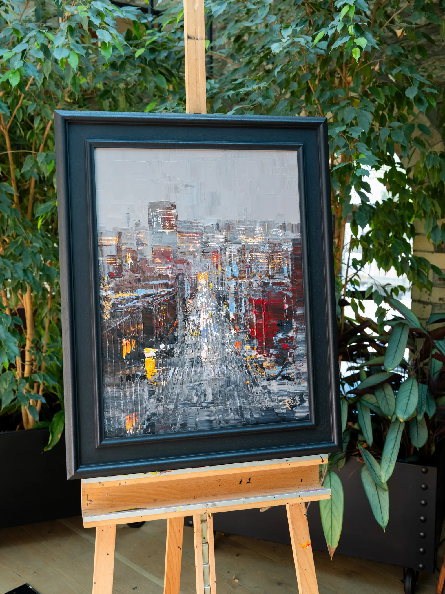 Midtown Silver, an original painting by Paul Kenton, shown in a dark wooden frame on a wooden easel among leafy plants. Thick silver and grey brushwork depicts a city street lined with glowing windows, with touches of red and amber.