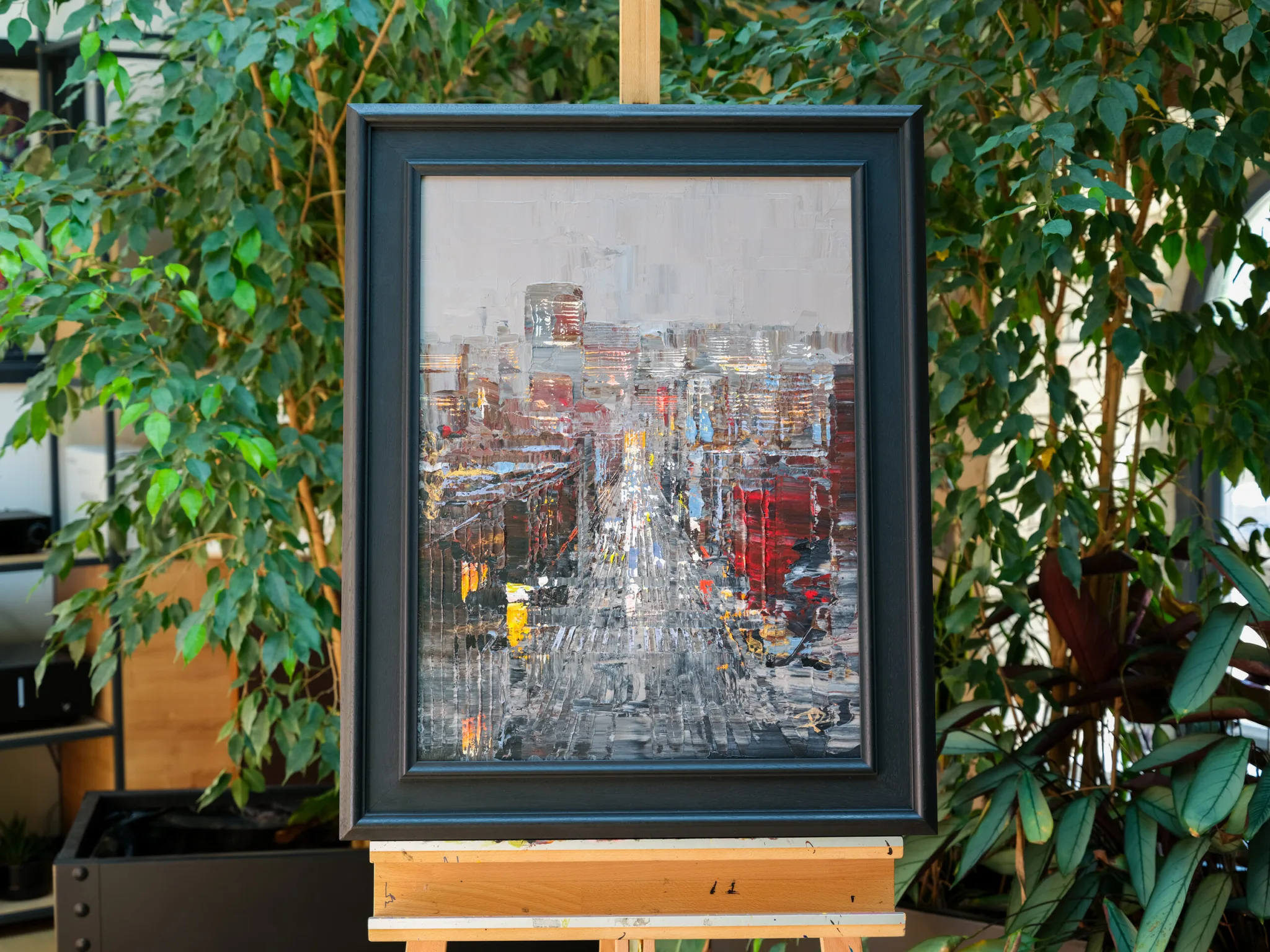 Midtown Silver, an original painting by Paul Kenton, shown in a dark wooden frame on a easel among green indoor plants. A textured city street scene in silver, grey and red, with glowing windows and towers.