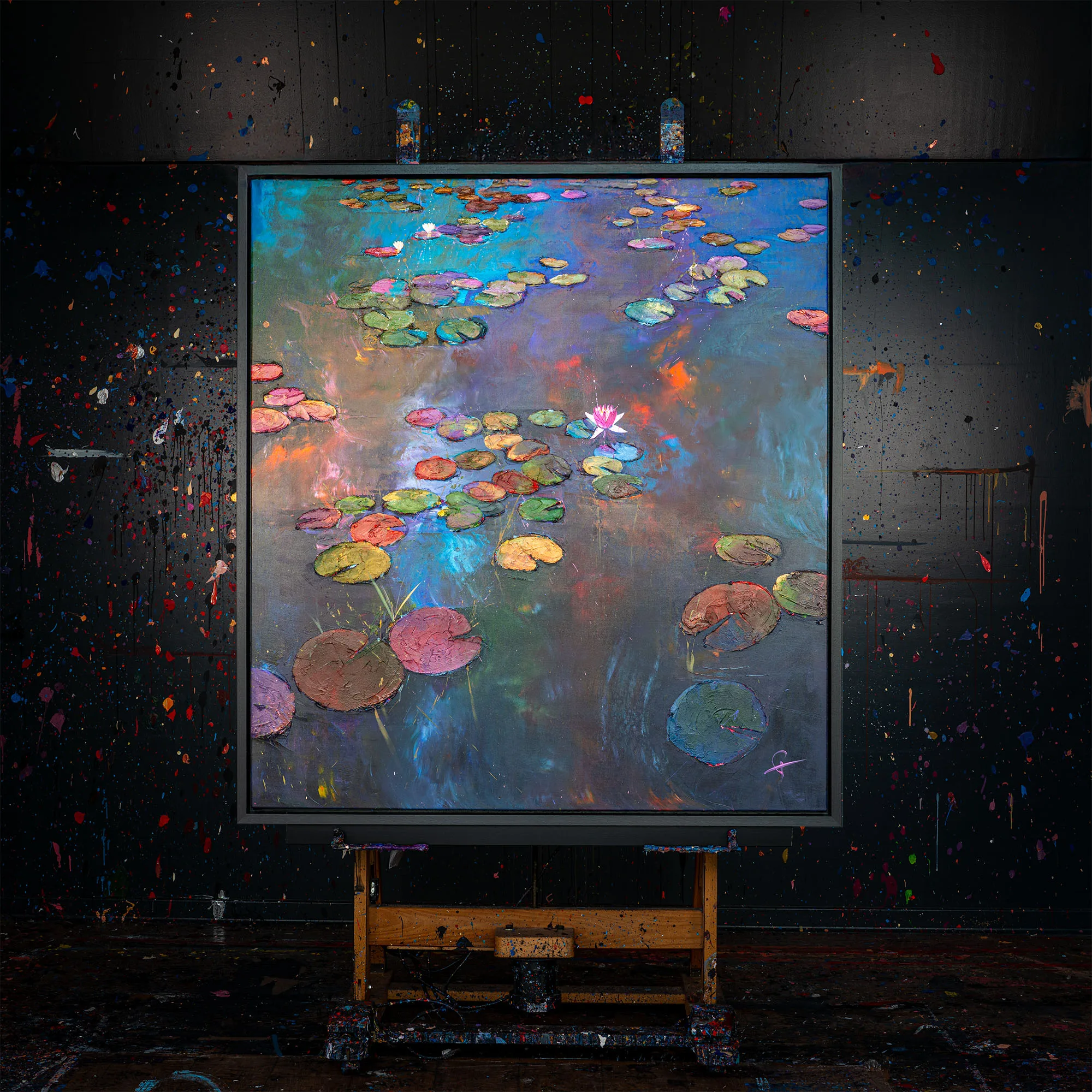 Nocturne Blossoms, an original mixed media painting by Paul Kenton, hangs on a wooden easel in a dark studio spattered with paint. Lily pads and a pink water lily float on a shimmering blue, violet and orange pond.