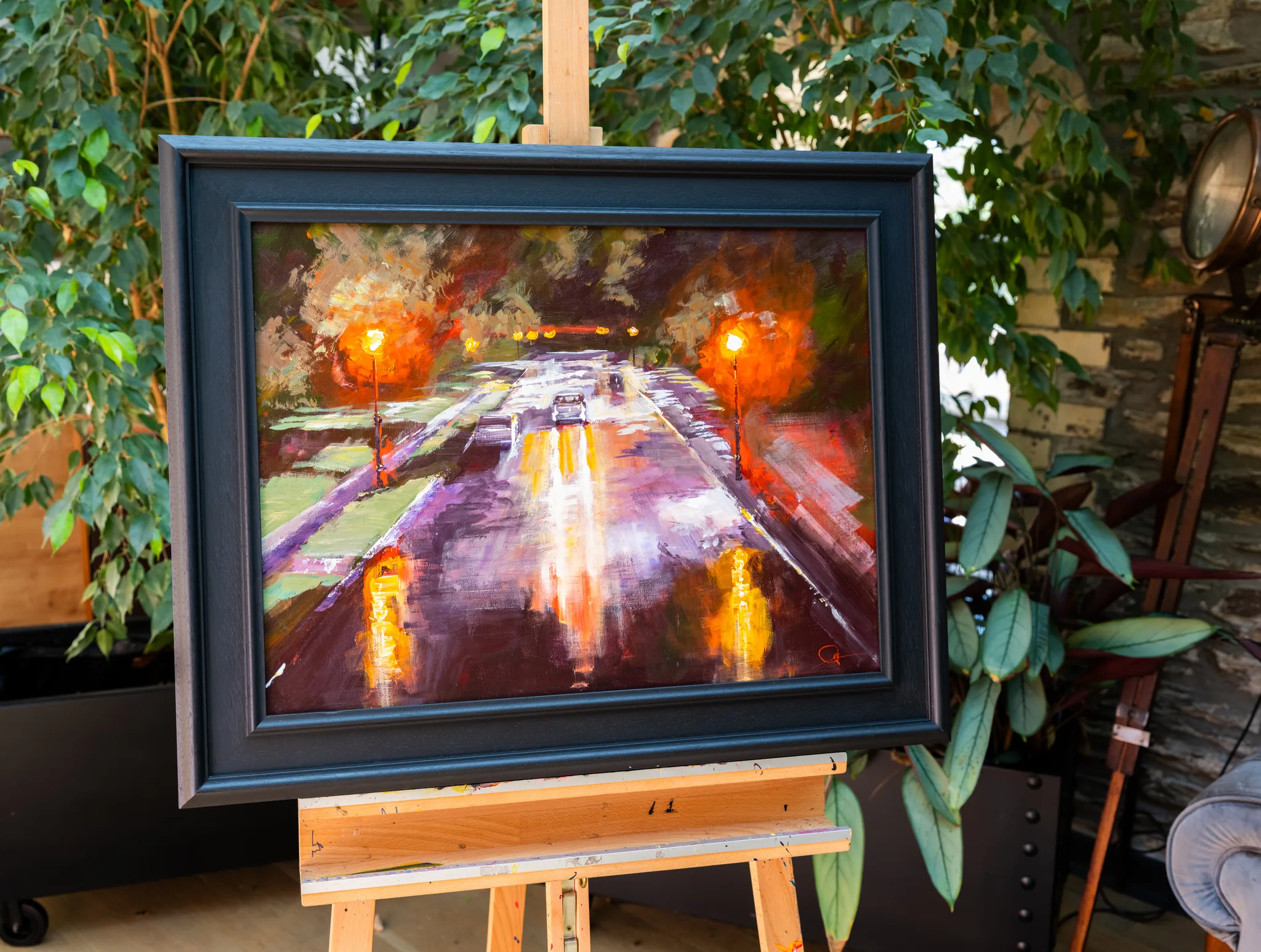 Night Drive, an original painting by Paul Kenton, hangs on a wooden easel in a leafy courtyard. Glowing street lamps and warm reflections light a wet city road at night.