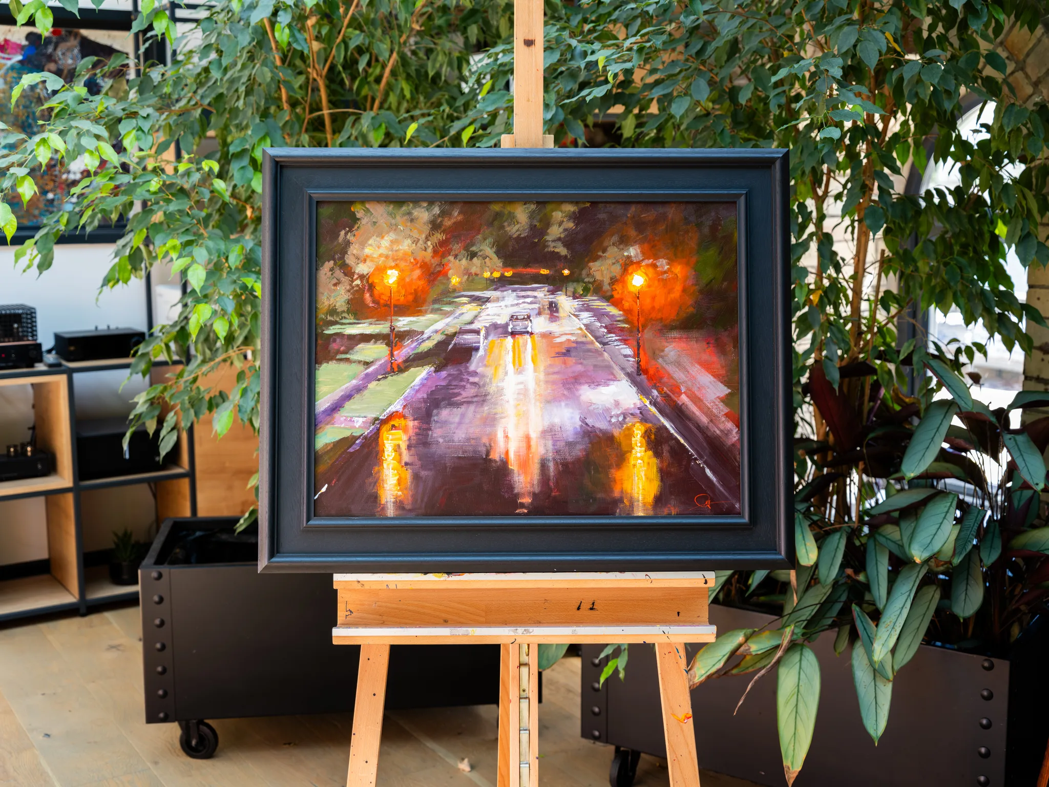 Night Drive, an original painting by Paul Kenton, in a dark frame on a wooden easel among indoor plants. It shows a rain-slick city street at night, lit by glowing orange streetlamps and reflections.