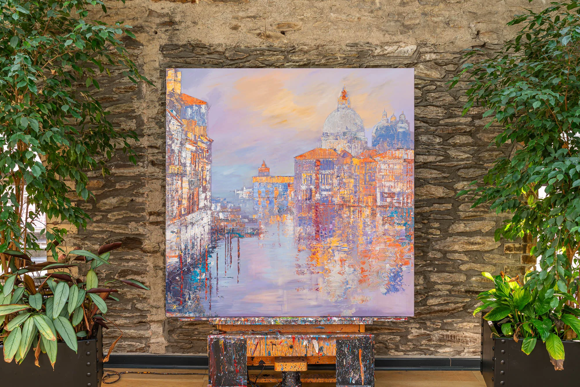 Opalescent Venice, an original painting by Paul Kenton hung on a stone wall between leafy plants, showing a glowing Venetian canal at dusk with a domed church, warm orange and lilac tones reflected in the water.