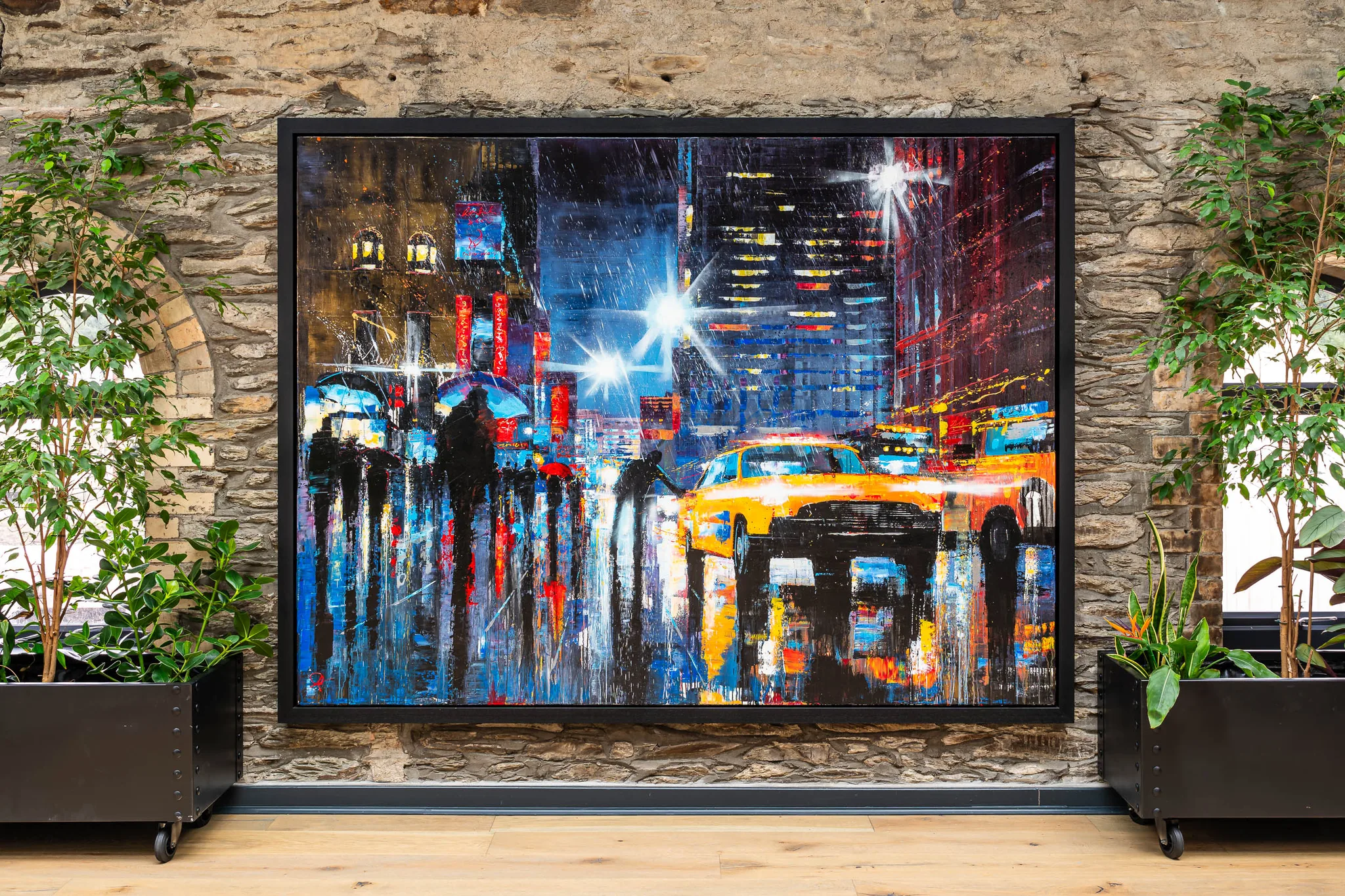 Taxi Dash, an original painting by Paul Kenton hung in a black frame on a stone wall between potted plants: a rain-soaked New York street at night, with a yellow taxi, umbrellas and glowing blue and red reflections.