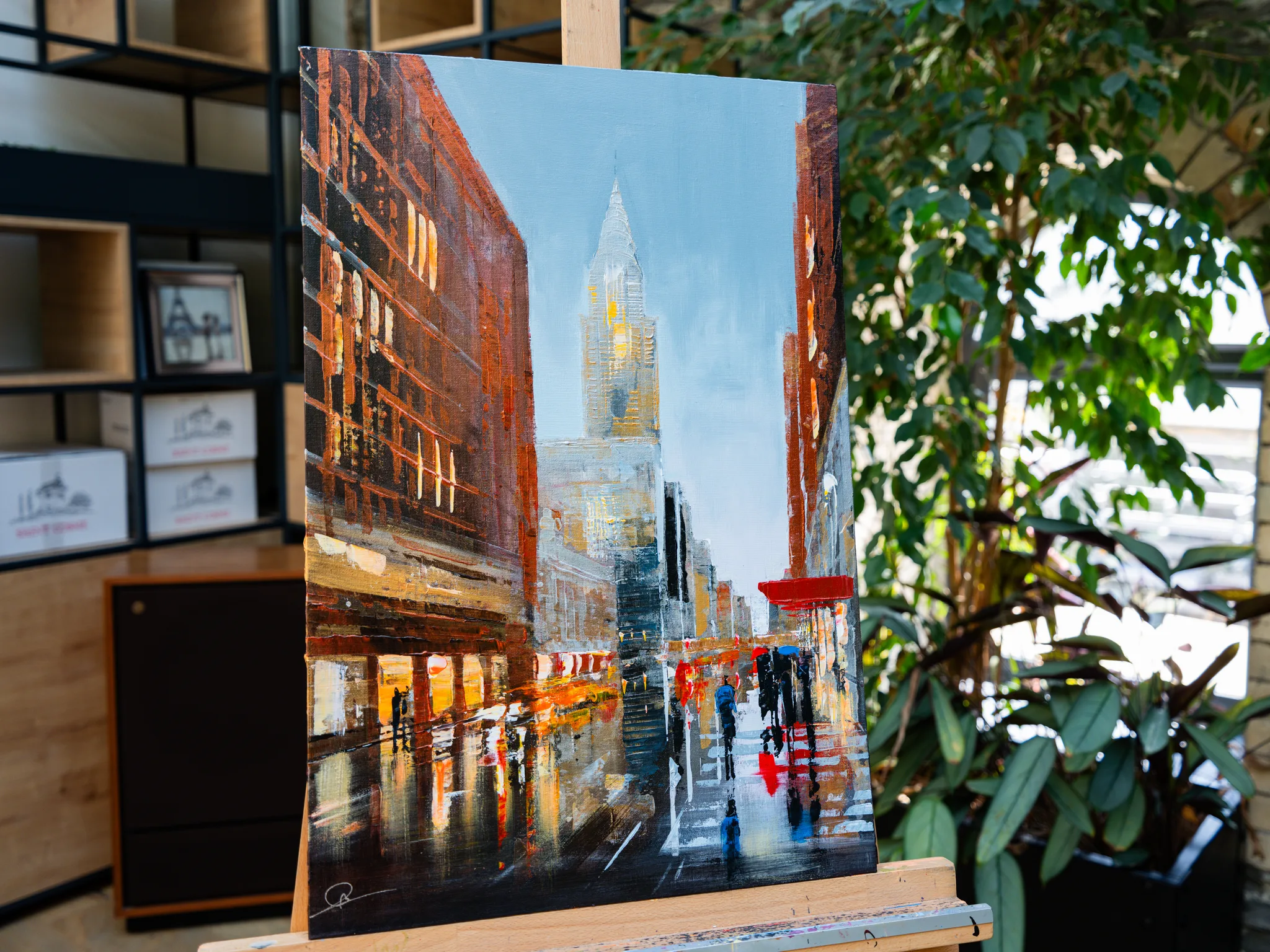 Under the Chrysler, an original painting by Paul Kenton, shown on an easel in a bright room: a rain-wet New York street of brick buildings, with a tall art deco tower glowing gold under a pale sky.