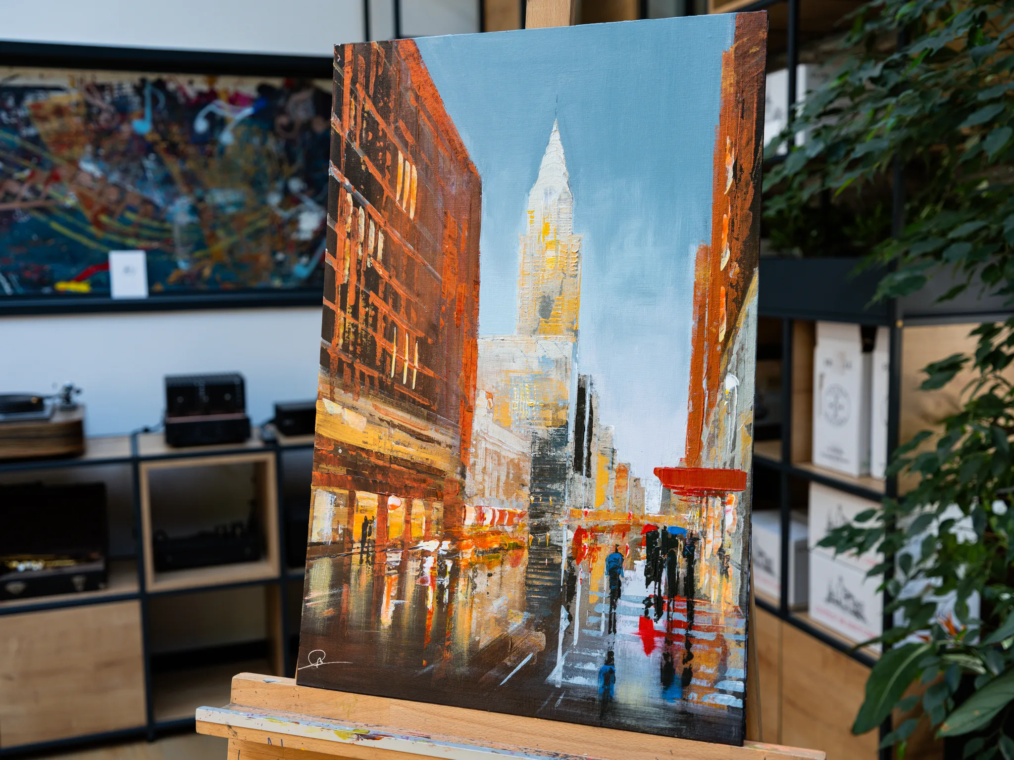 Under the Chrysler, an original painting by Paul Kenton, on an easel in a showroom. A rain-slicked New York street rises toward a gilded, spire-topped tower beneath a pale blue sky, framed by warm orange brick buildings.