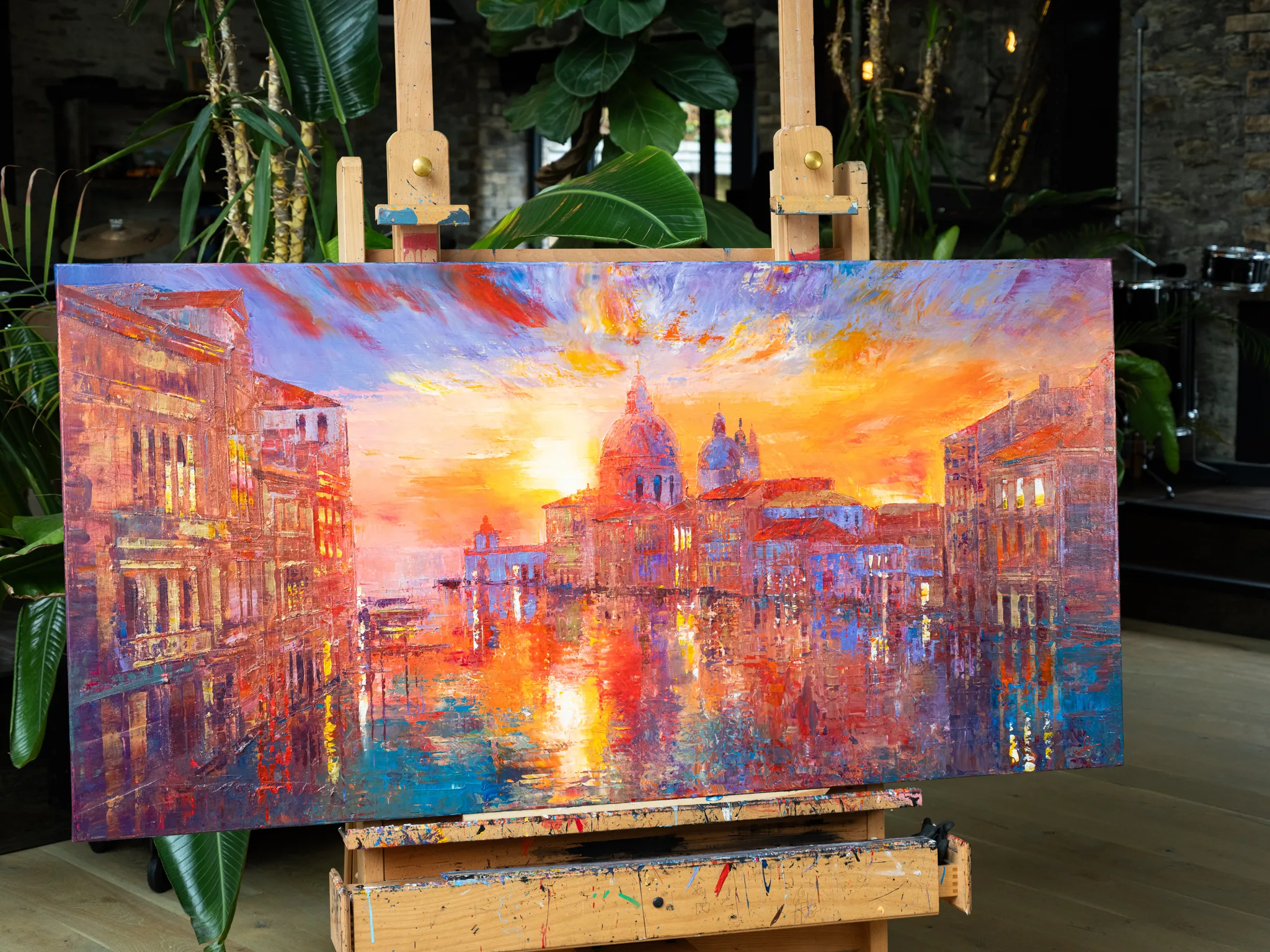 Venice Ablaze, an original painting by Paul Kenton, on a wooden easel among indoor plants, shows a fiery sunset over Venetian domes and canal.