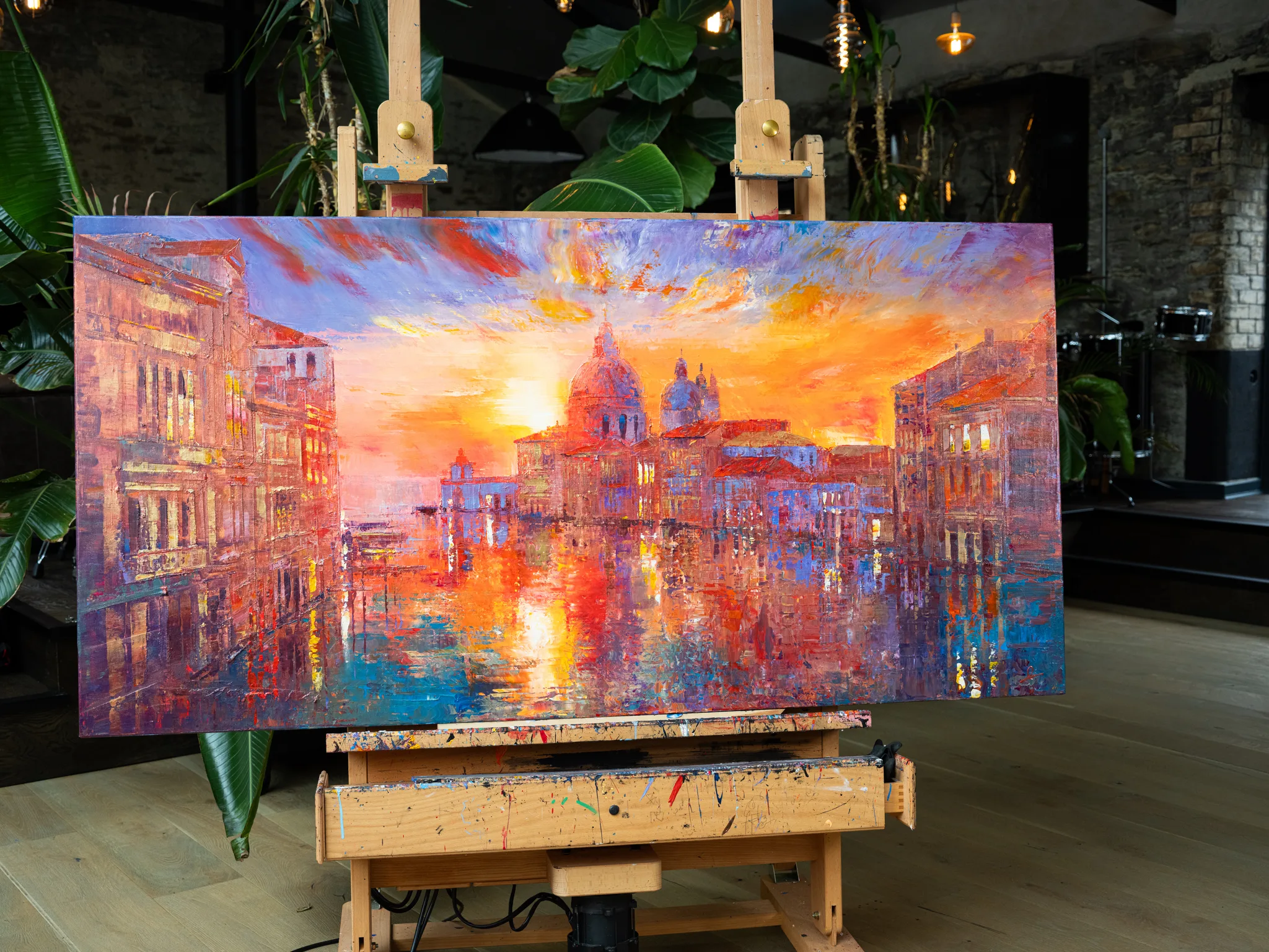 Venice Ablaze, an original painting by Paul Kenton, on an easel in a room with plants and exposed brick. Fiery orange and violet sunset lights a canal lined with palazzos and domed churches.