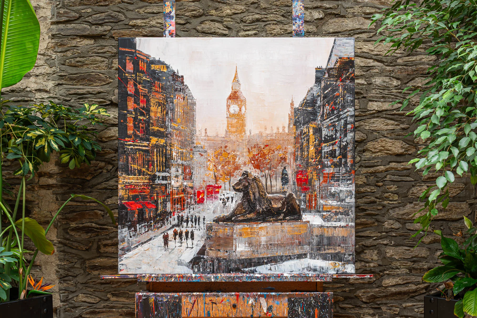 Westminster Watch, an original painting by Paul Kenton shown on an easel against a stone wall: a textured London street scene with a bronze lion in the foreground and Big Ben glowing in warm amber light beyond.