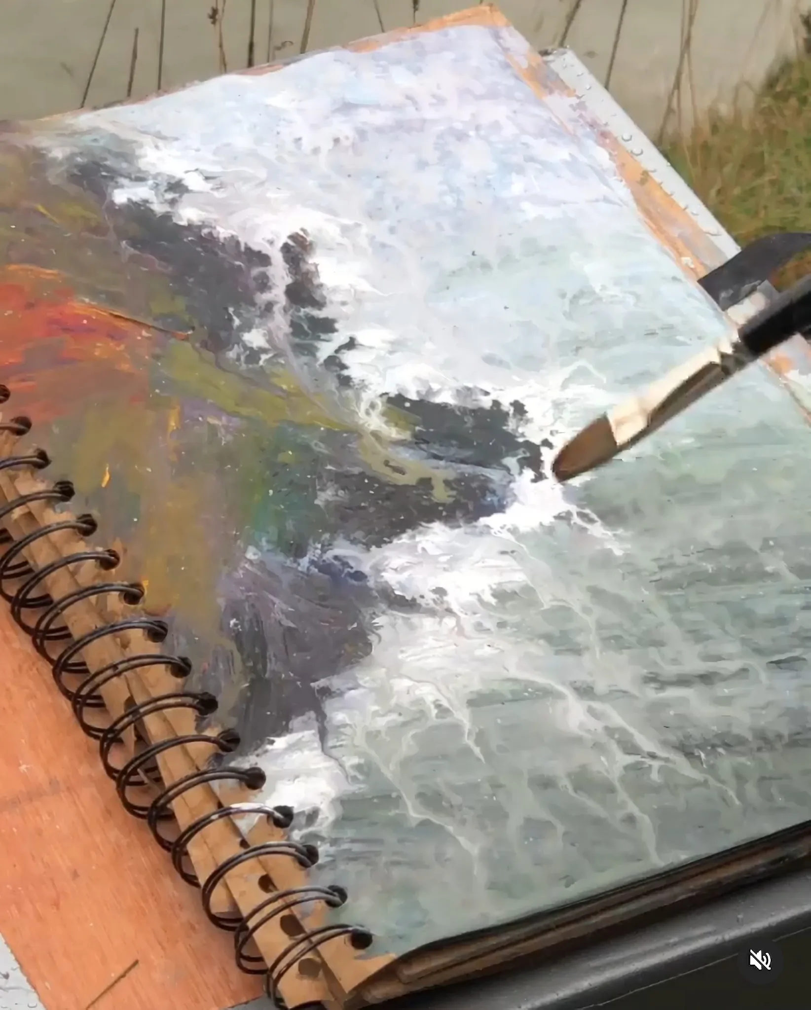 Against the Elements, an original gouache by Paul Kenton, shown on a wooden board outdoors, a brush over grey-blue waves and dark rocks.
