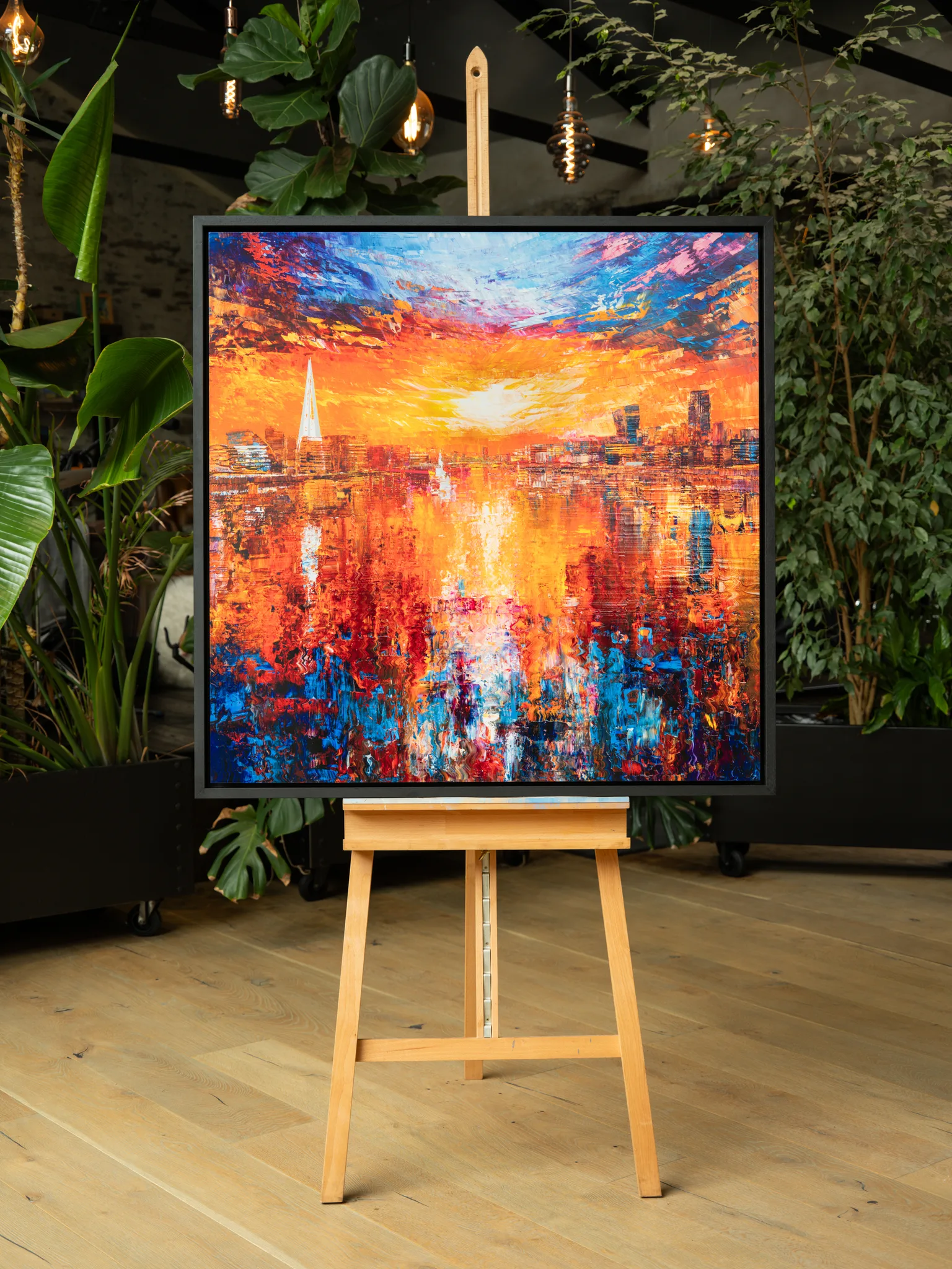 Currents of Light, a limited edition print by Paul Kenton, framed in black on a wooden easel among indoor plants. A bold sunset over a London skyline burns orange and red, reflected in rippling blue and red water.