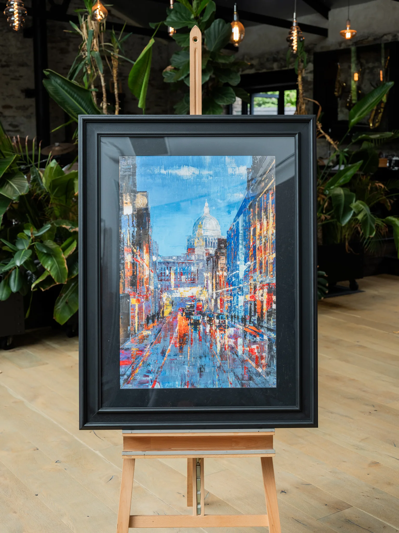 Cathedral of Light, a limited edition print by Paul Kenton, hangs in a black frame on a wooden easel in a plant-filled room, showing a blue-toned London street at dusk with St Paul's dome and warm lights.