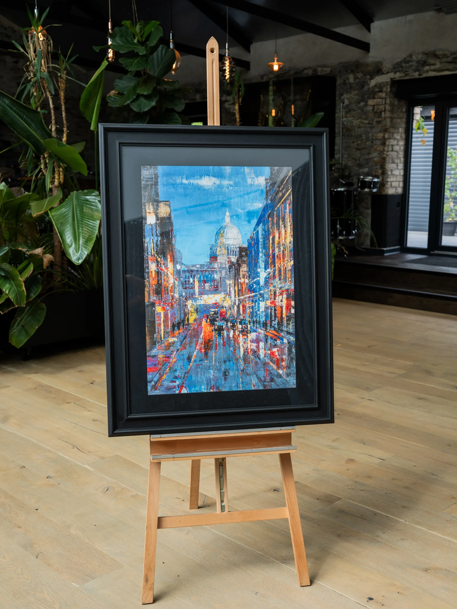 Cathedral of Light, a limited edition print by Paul Kenton, displayed in a black frame on a wooden easel in a room with plants and exposed brick. It shows a rain-slicked London street at dusk, lit by warm windows, with a domed cathedral glowing at the end under blue sky.