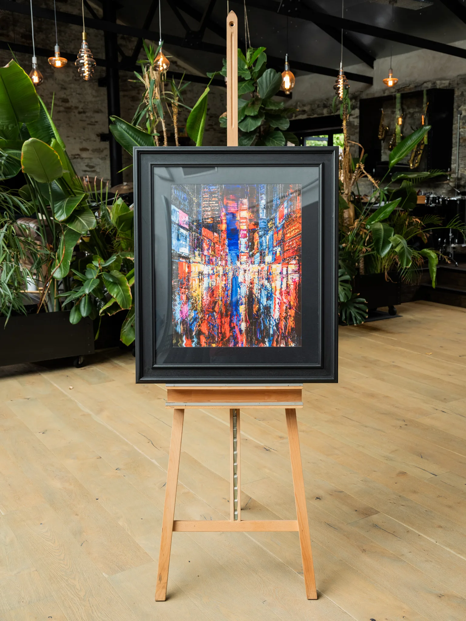 Downtown Dazzle, a limited edition print by Paul Kenton, hangs in a black frame on a wooden easel in a plant-filled room, showing a New York street of glowing red, orange and blue lights.