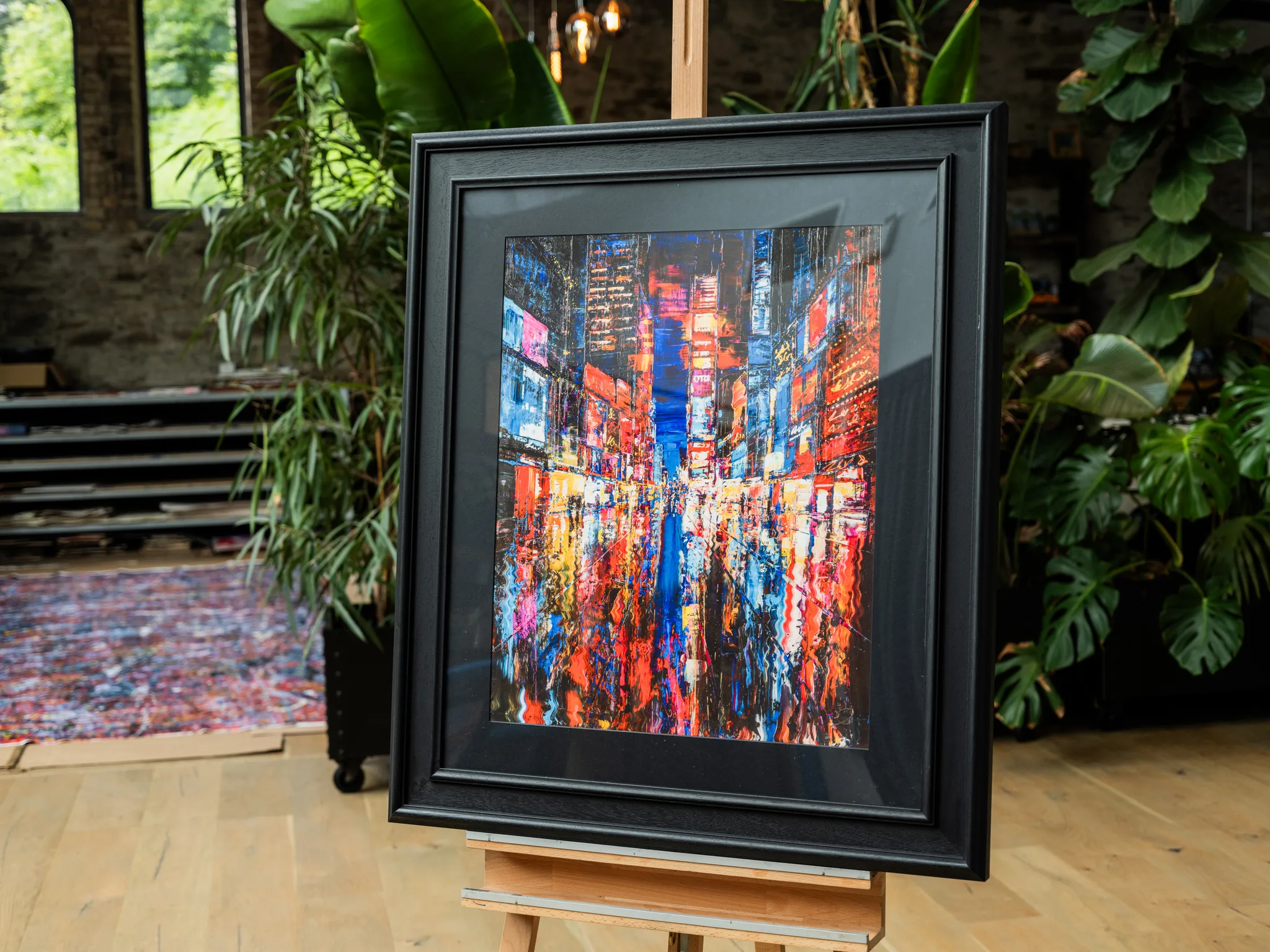 Downtown Dazzle, a limited edition print by Paul Kenton, shown in a black frame on an easel in a plant-filled room, depicting a glowing New York street of red and blue neon.