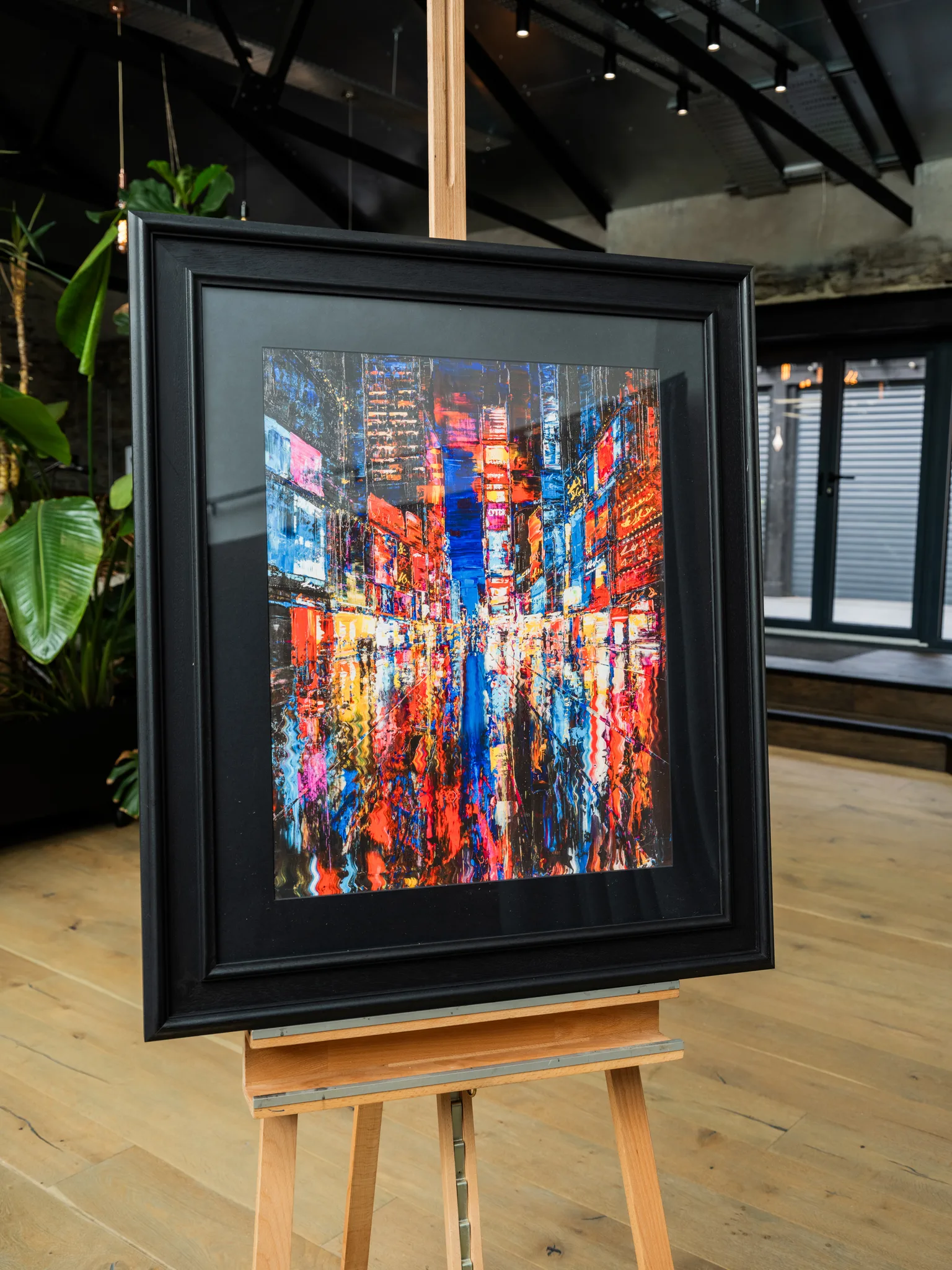 Downtown Dazzle, a limited edition print by Paul Kenton, hangs in a black frame on a wooden easel in a bright room with plants. It shows a New York street at night, glowing with red, orange and blue lights and reflected on wet pavement.