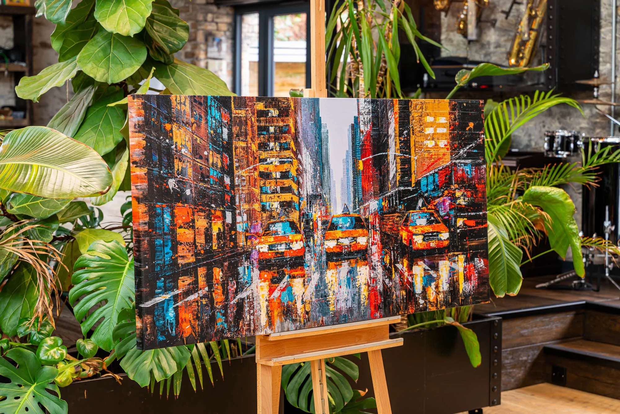 Downtown Reflections, a limited edition canvas print by Paul Kenton, on a wooden easel among large green plants; a bold, textured night street scene with glowing orange and blue lights and yellow taxis.